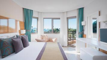 A hotel room with a large bed, a sofa, a desk, and a balcony with a view of palm trees and mountains.