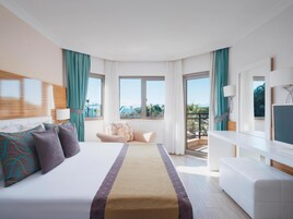 Een hotelkamer met een groot bed, een bank, een bureau en een balkon met uitzicht op palmbomen en bergen.