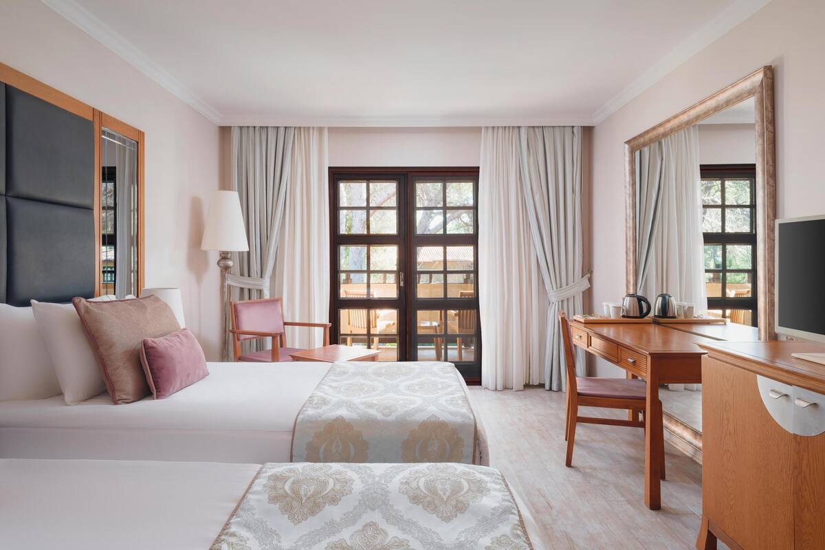A hotel room with two beds, a desk, a chair, a television, and large windows with curtains.