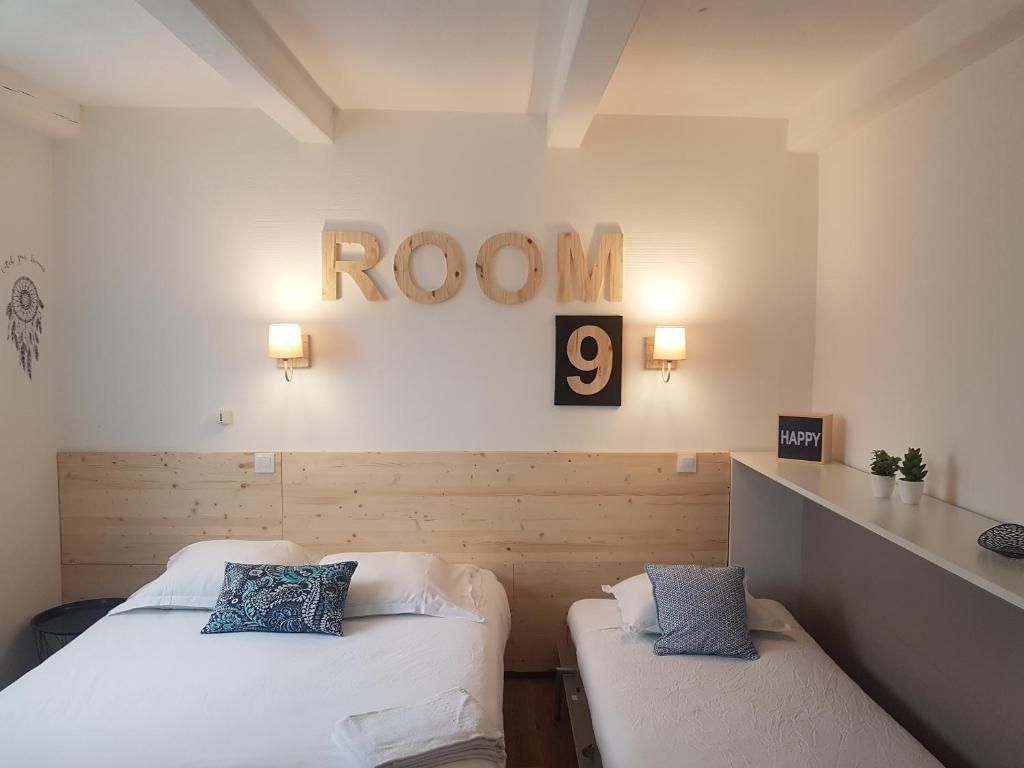 A hotel room with two beds, a wooden headboard, a wall-mounted sign indicating 'ROOM 9', and a small shelf with a 'HAPPY' sign.
