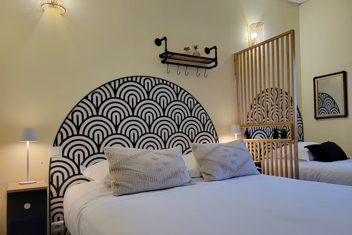 A hotel room with a large, circular, black and white patterned headboard, two bedside tables with lamps, a bed with white bedding and two decorative pillows, a wooden slat partition, and a framed artwork on the wall.