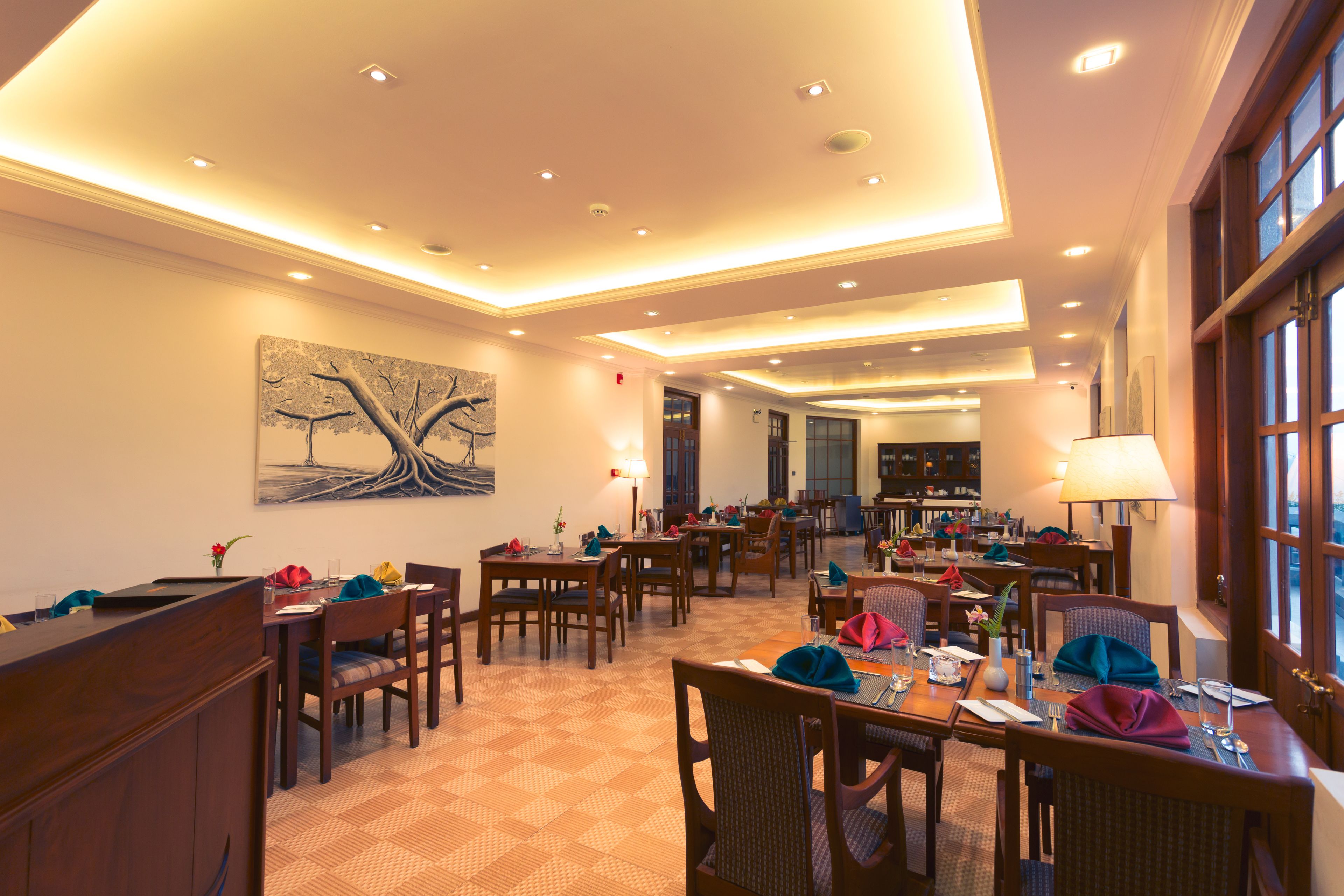 A hotel dining area with multiple tables and chairs, a reception desk, and a large painting on the wall.