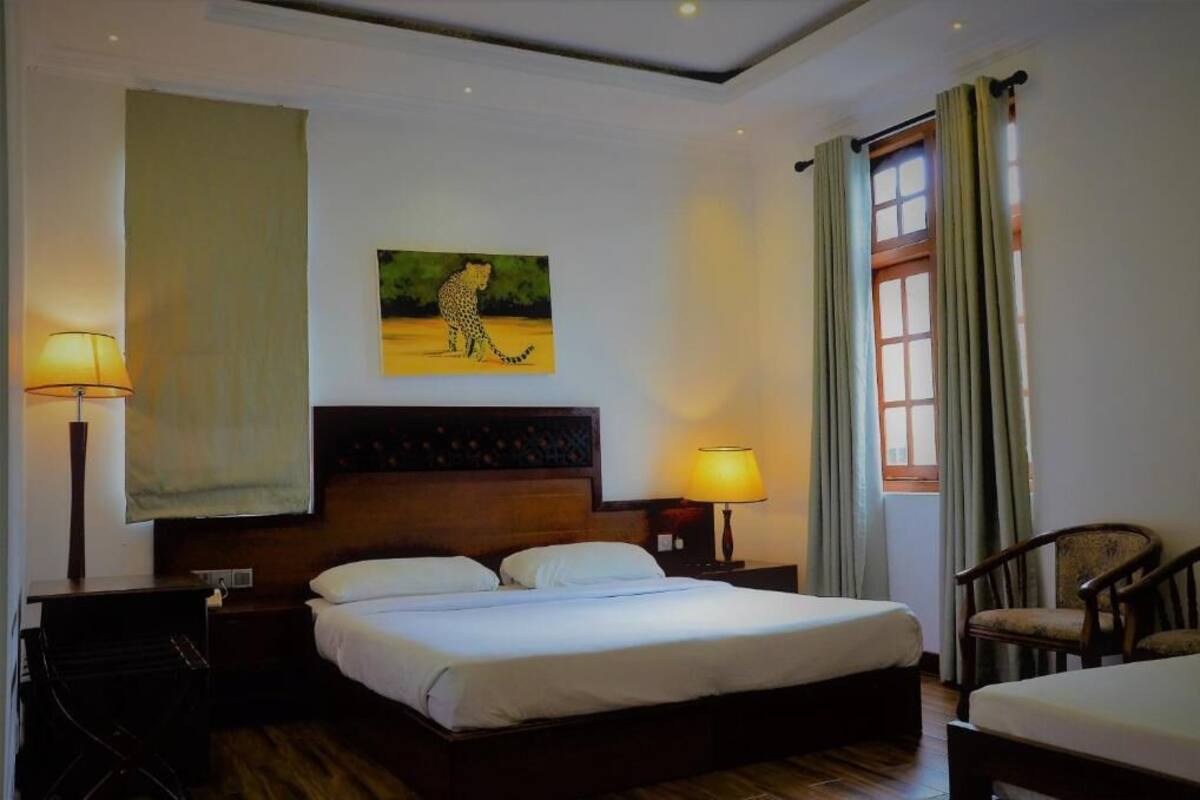 A hotel room with a large bed, two bedside lamps, a painting of a leopard, a window with curtains, and a chair.