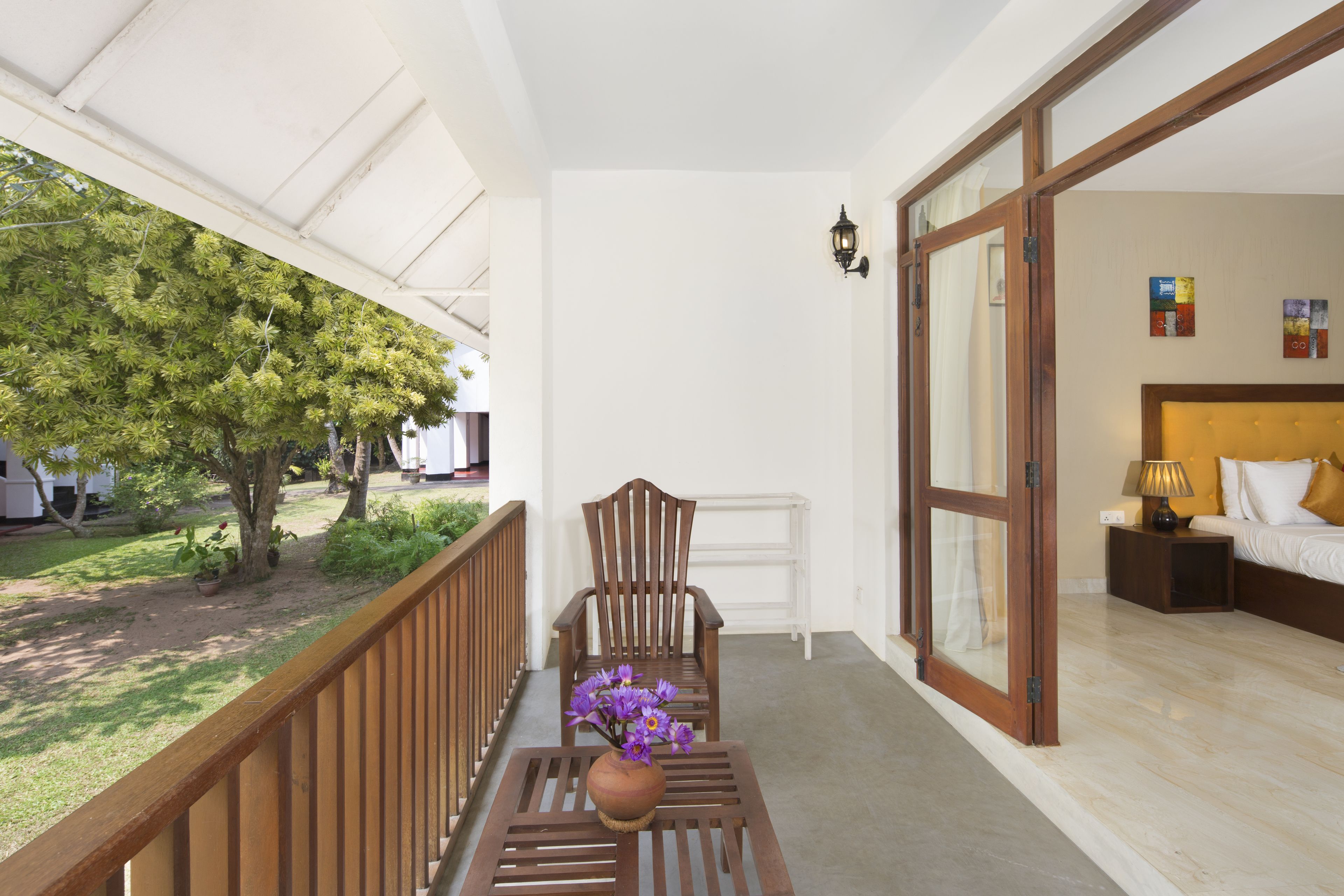 A balcony with a wooden chair and a vase of flowers, a bed with a nightstand, and a view of a tree and grass.