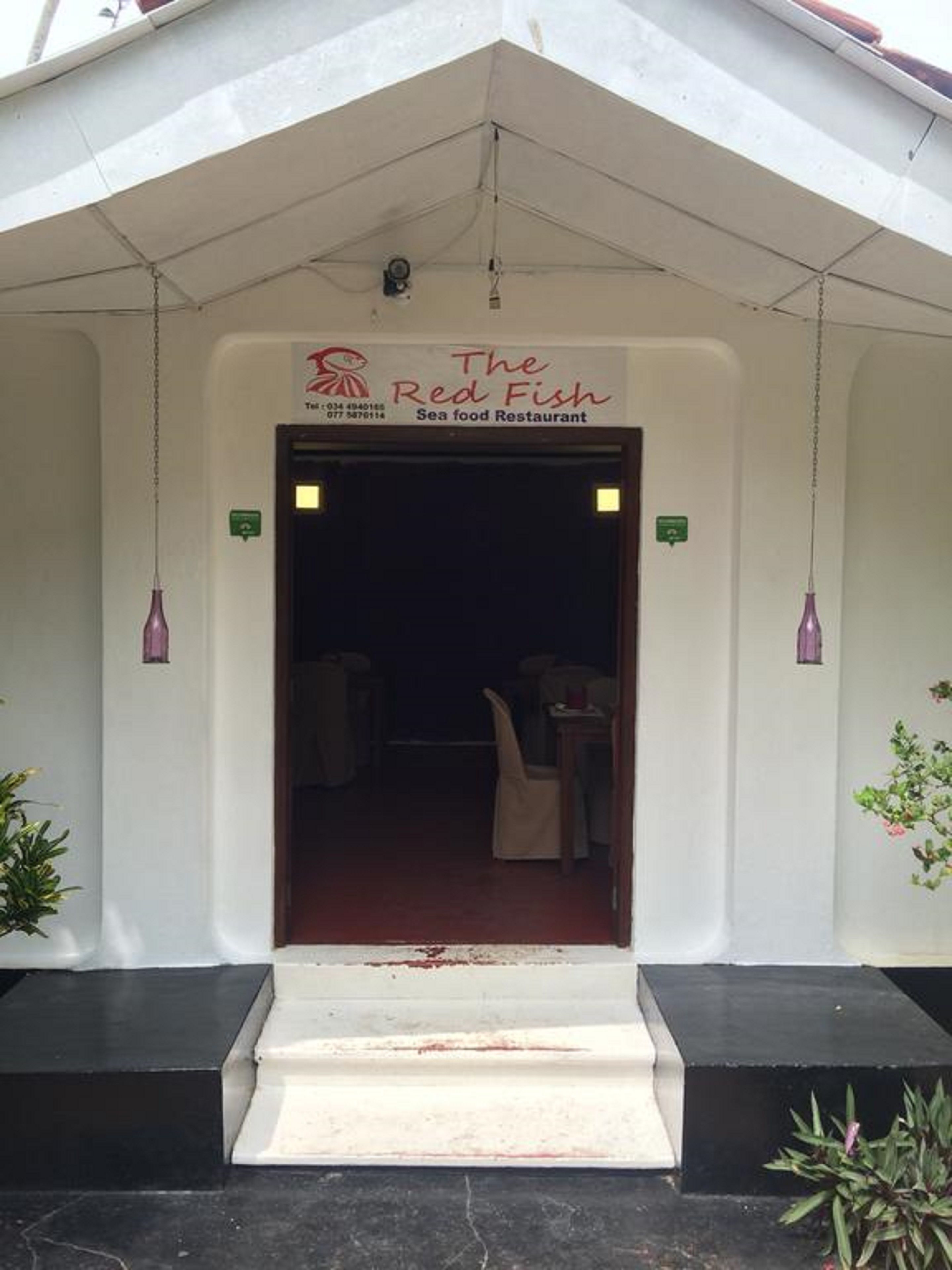 The entrance to The Red Fish Sea food Restaurant with a simple white facade, a dark interior visible through the doorway, and a small outdoor seating area with chairs and tables.
