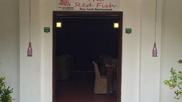 The entrance to The Red Fish Sea food Restaurant with a simple white facade, a dark interior visible through the doorway, and a small outdoor seating area with chairs and tables.