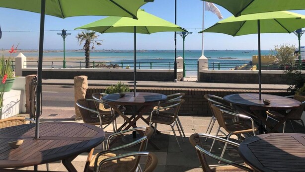 An outdoor seating area with tables and chairs, overlooking a beach. - De L'Etang Jersey (St. Helier)