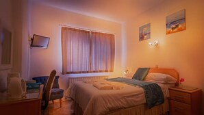 A bedroom with a bed, a desk, a chair, a window with curtains, and wall-mounted lights. - De L'Etang Jersey (St. Helier)