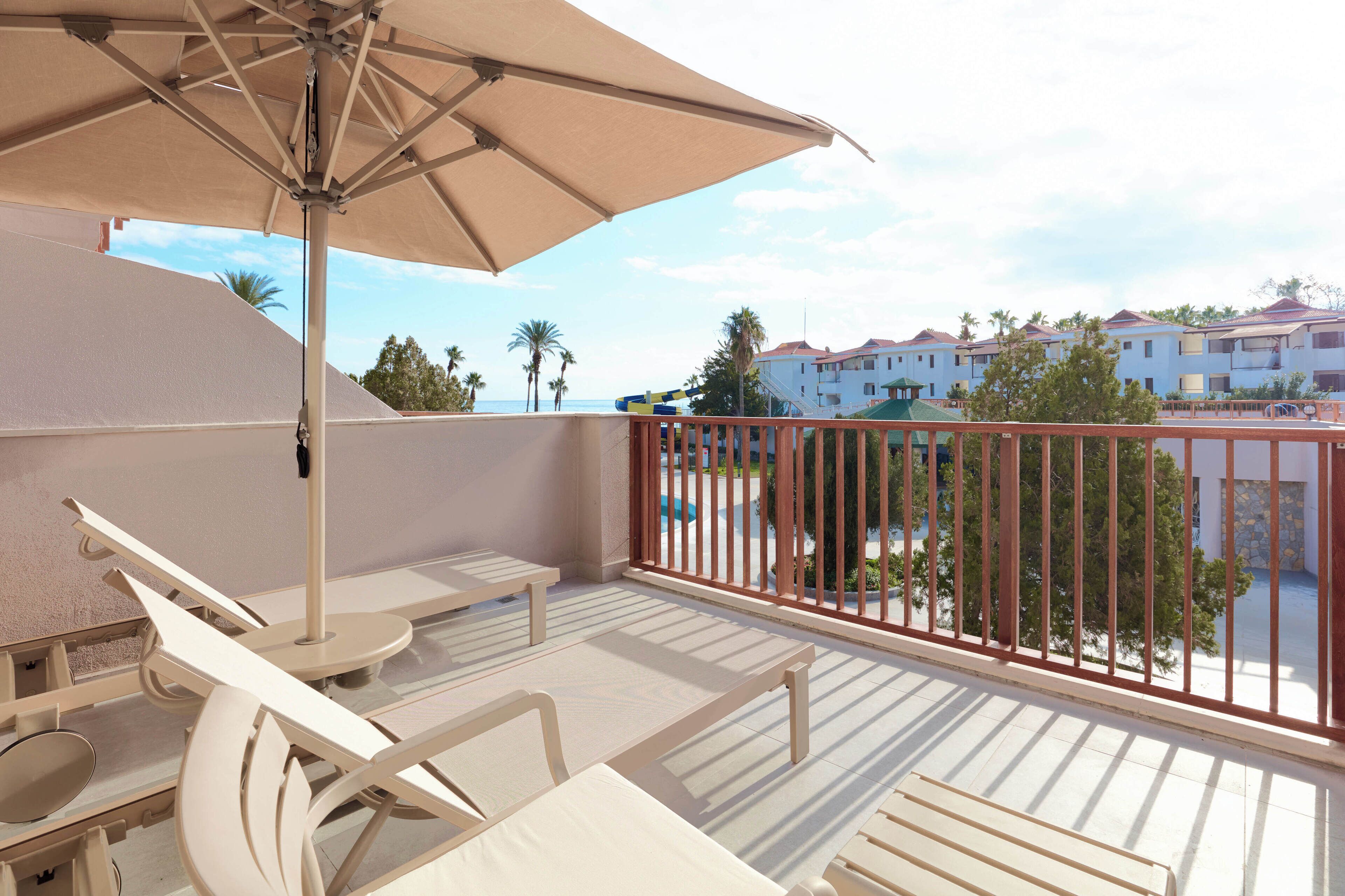 En balkon med hvide liggestole og en stor parasol, med udsigt over en pool og bygninger.