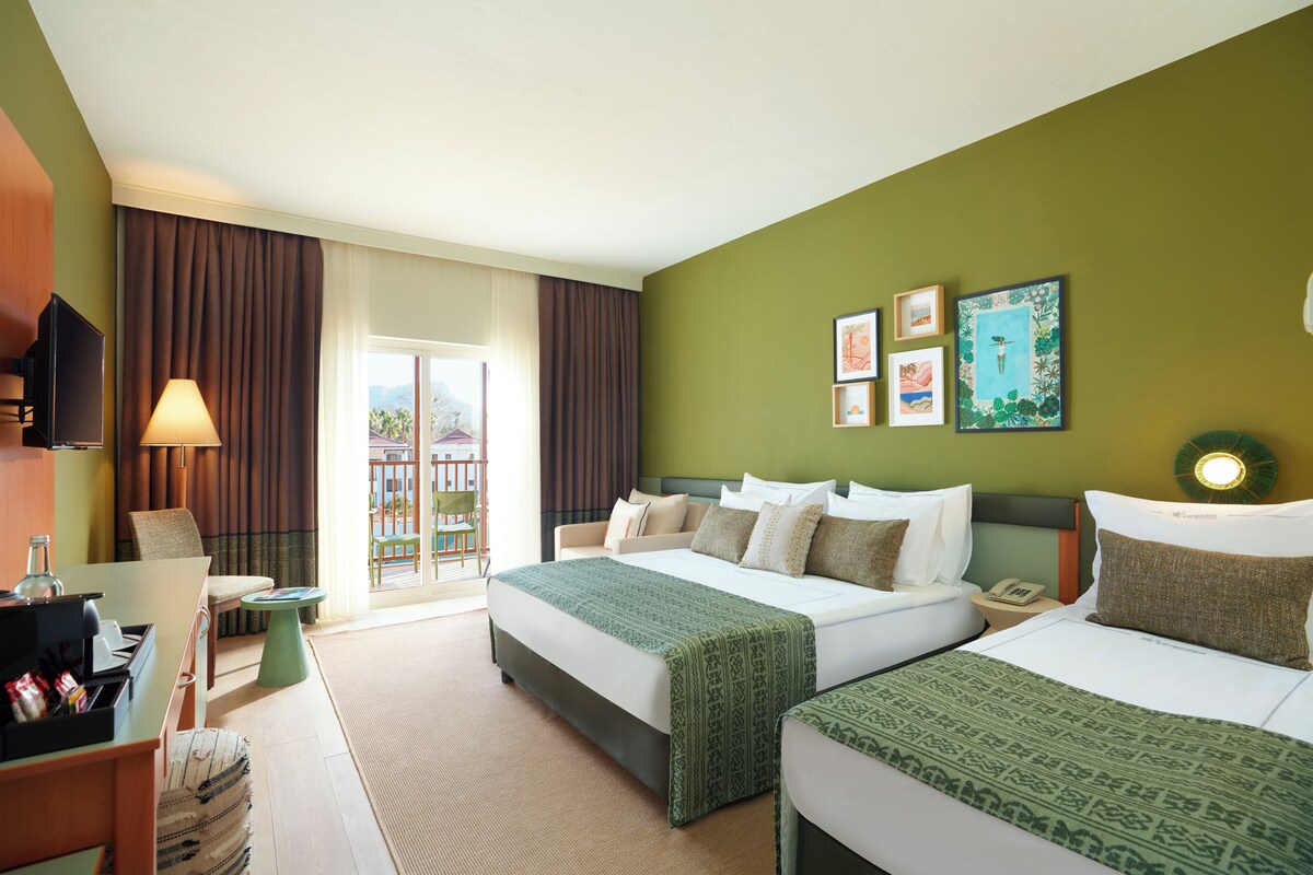 Habitación de hotel con dos camas, un escritorio, una silla, una lámpara y un balcón con cortinas.