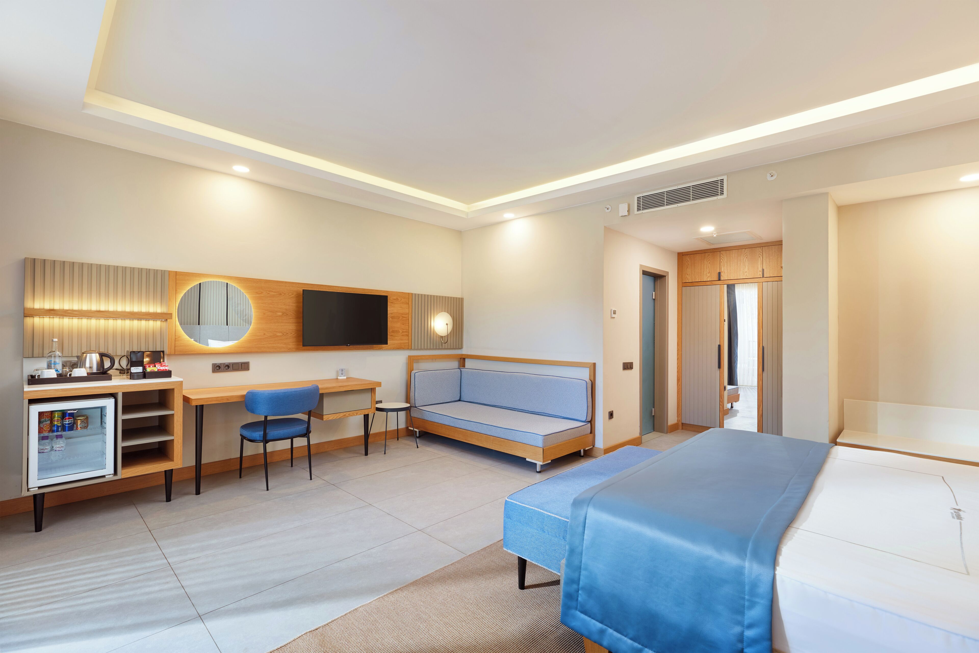 A modern hotel room with a bed, a sofa, a desk with a computer, a small fridge, and a television.