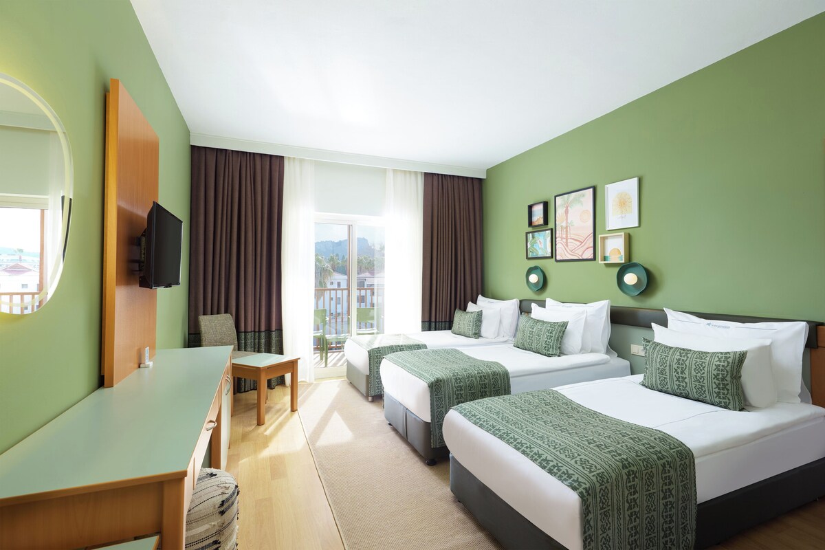 Habitación de hotel con dos camas, un escritorio, una silla, televisión y balcón con cortinas.