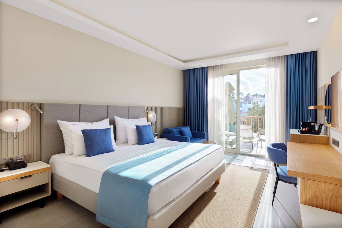 A modern hotel room with a large bed, a desk, and a balcony with a view.