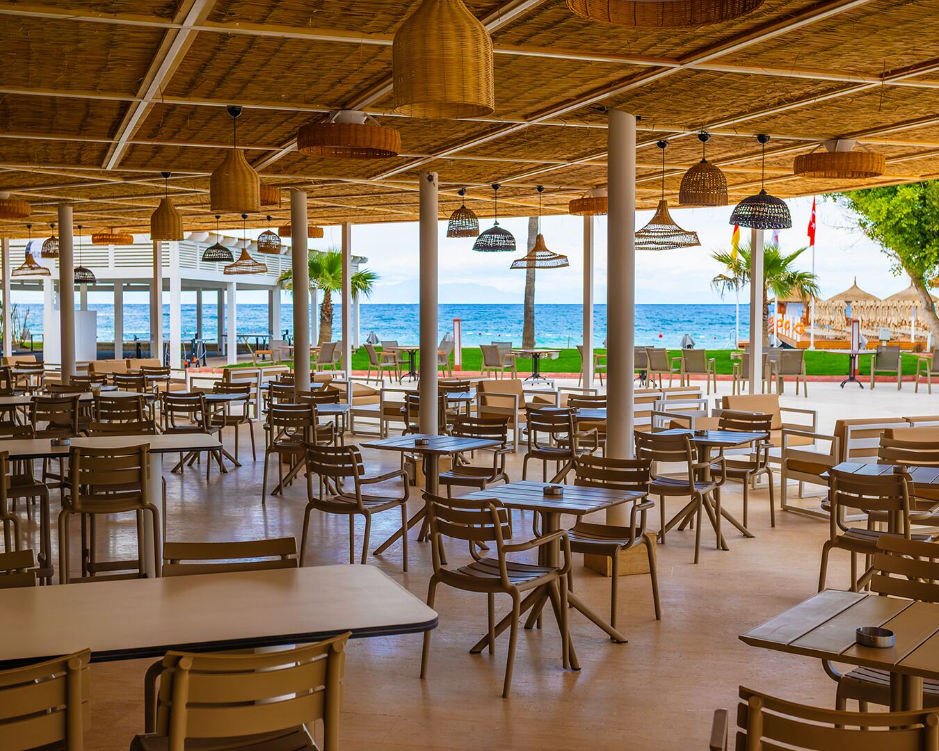 Un área de comedor al aire libre con mesas y sillas, vista al mar y techo de paja.