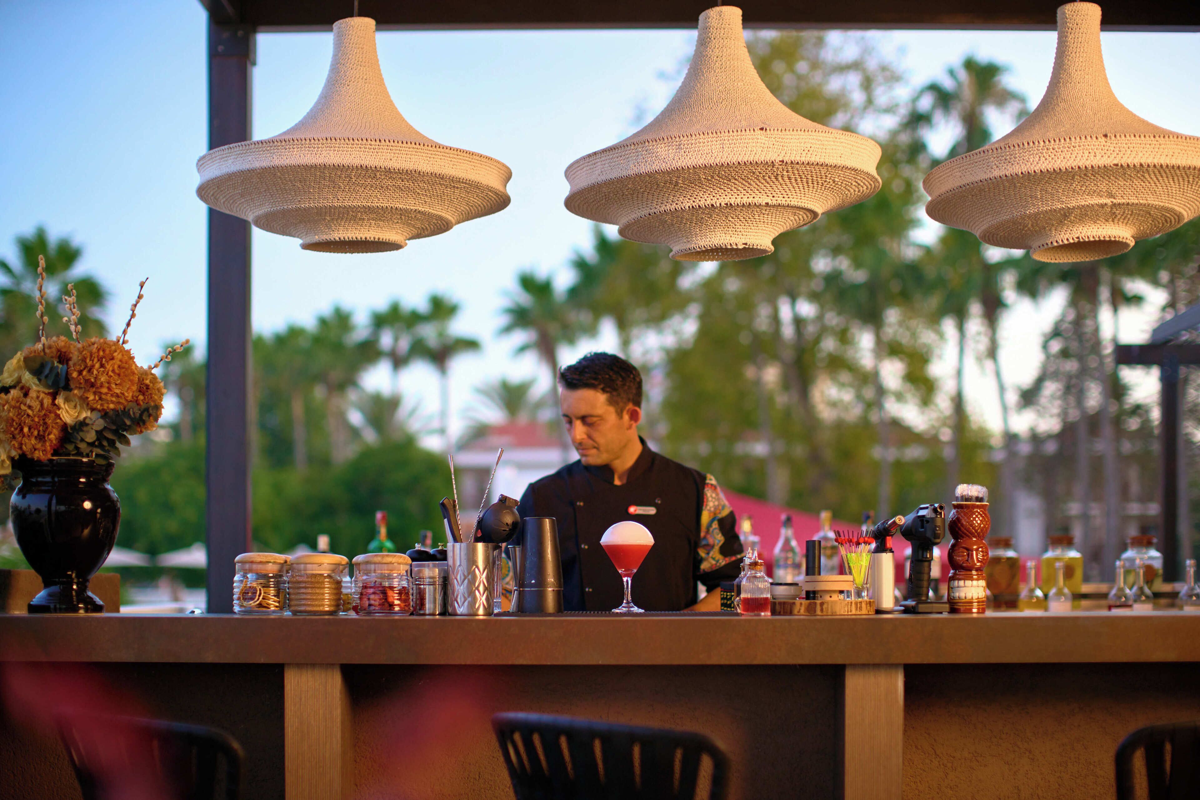 Uma área de bar com luminárias pendentes, um barman e diversos utensílios e garrafas de bar.