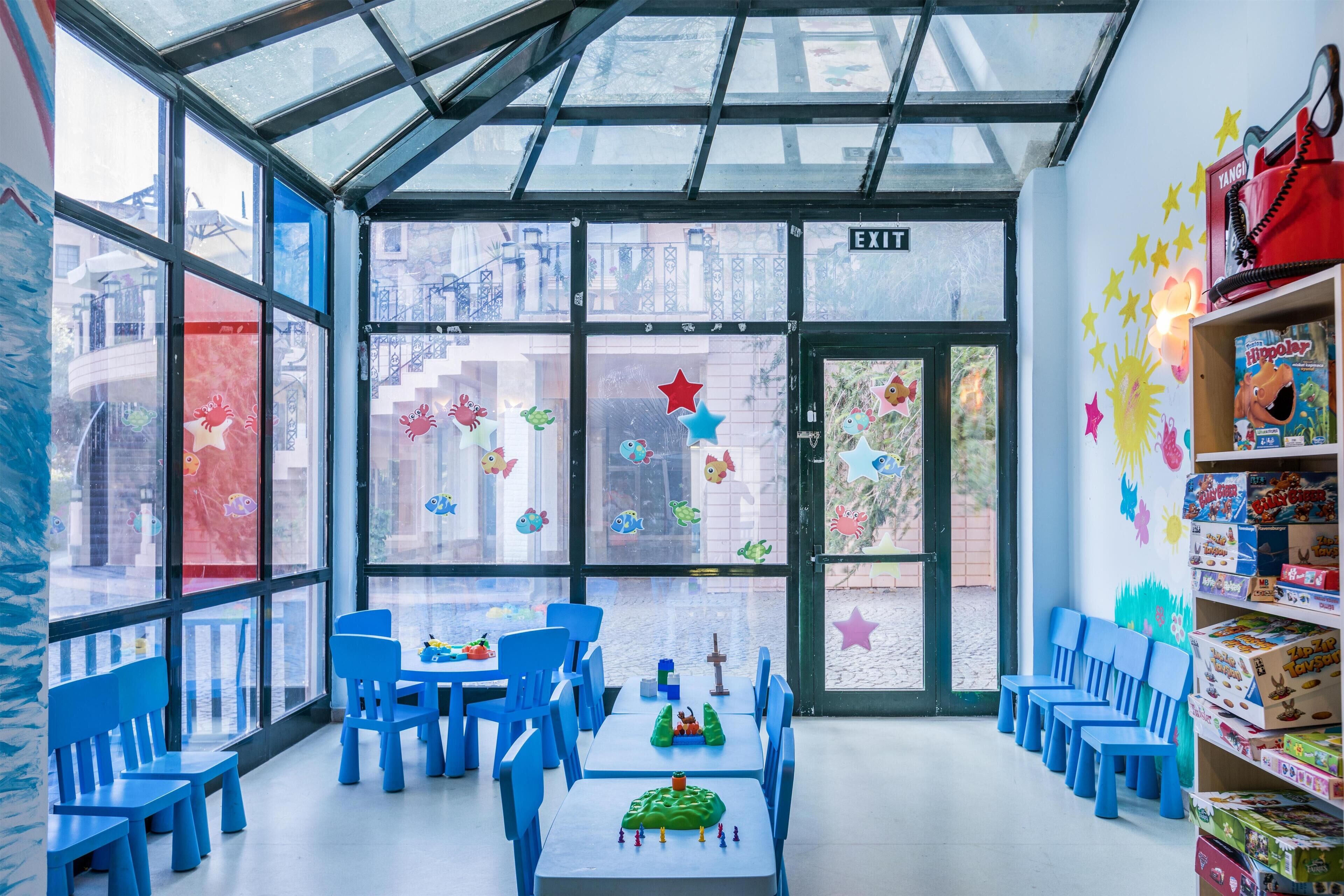 Zona de juegos infantiles con sillas y mesas azules, techo de cristal y pared decorada con estrellas de colores.