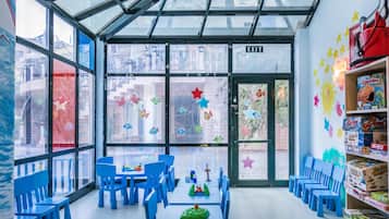 Zona de juegos infantiles con sillas y mesas azules, techo de cristal y pared decorada con estrellas de colores.
