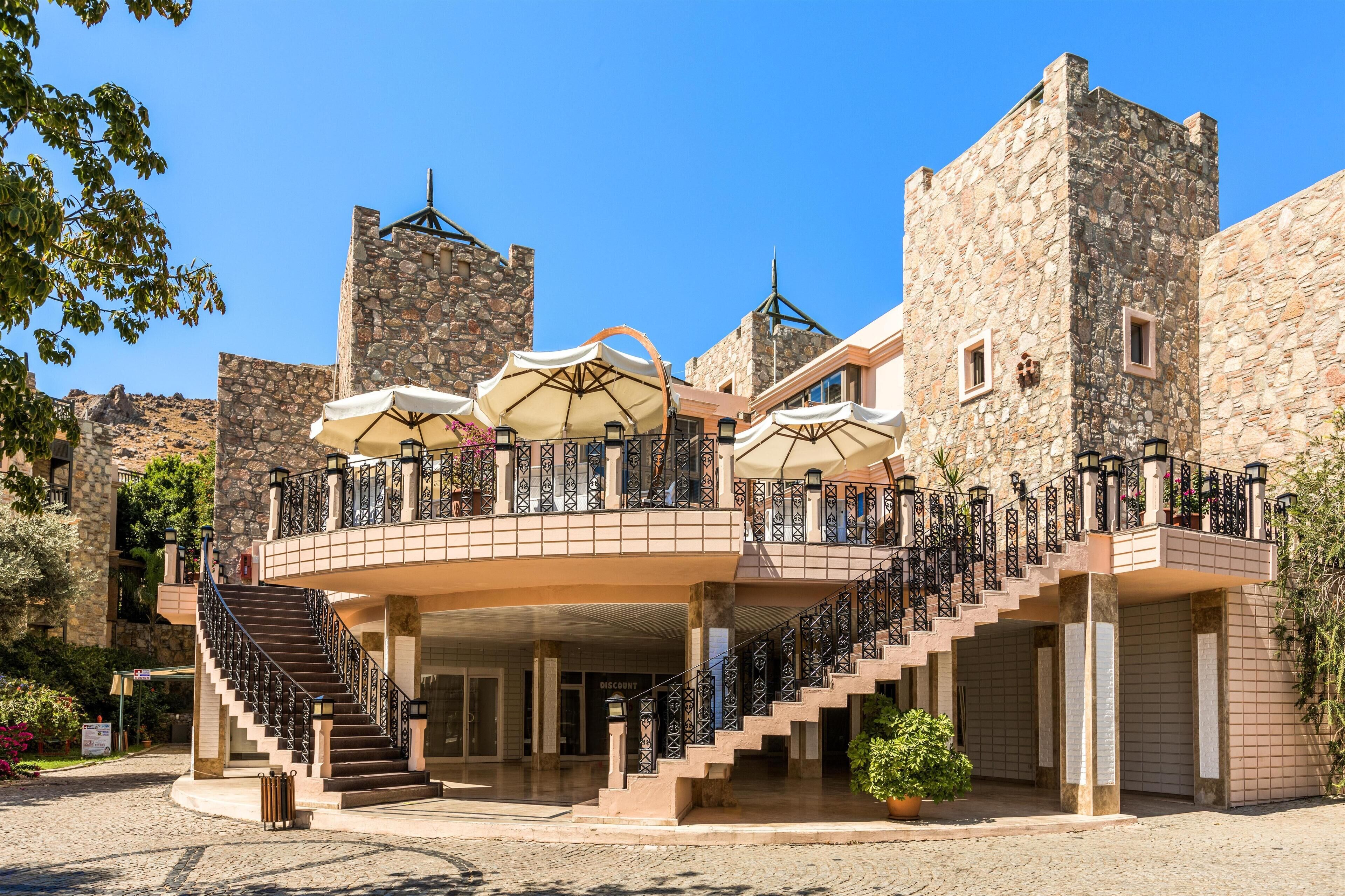 Un edificio storico con facciata in pietra, una scala centrale e posti a sedere all'aperto con ombrelloni.