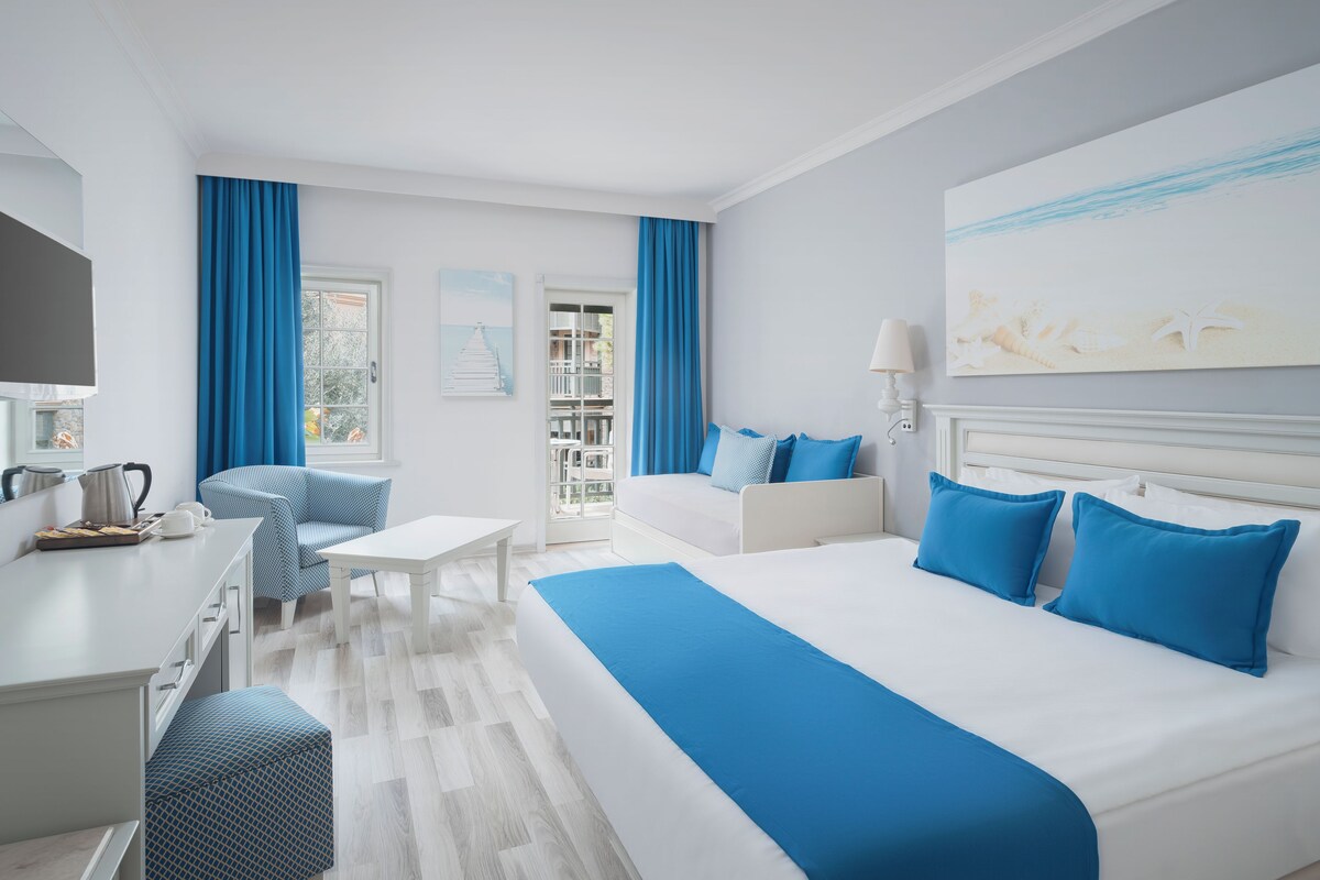 A modern hotel room with a large bed, a desk, a chair, and a balcony with blue curtains.