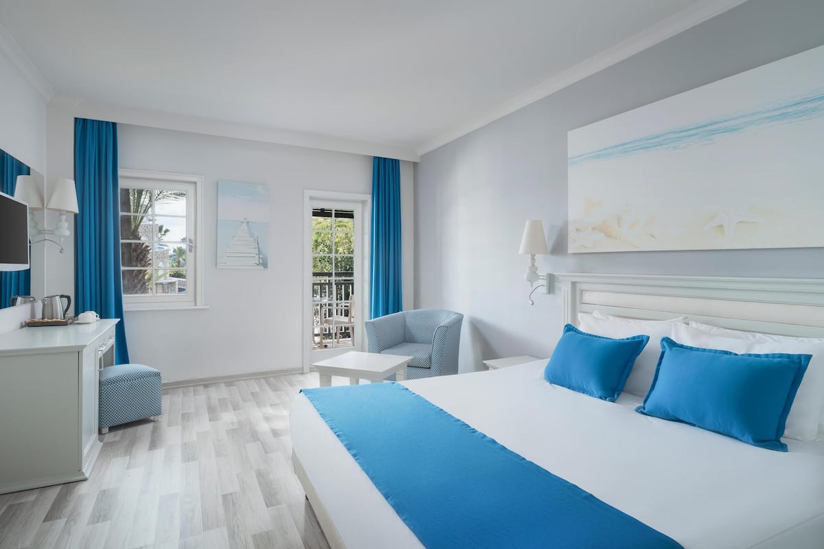A hotel room with a large bed, a desk, a chair, and a balcony with blue curtains.