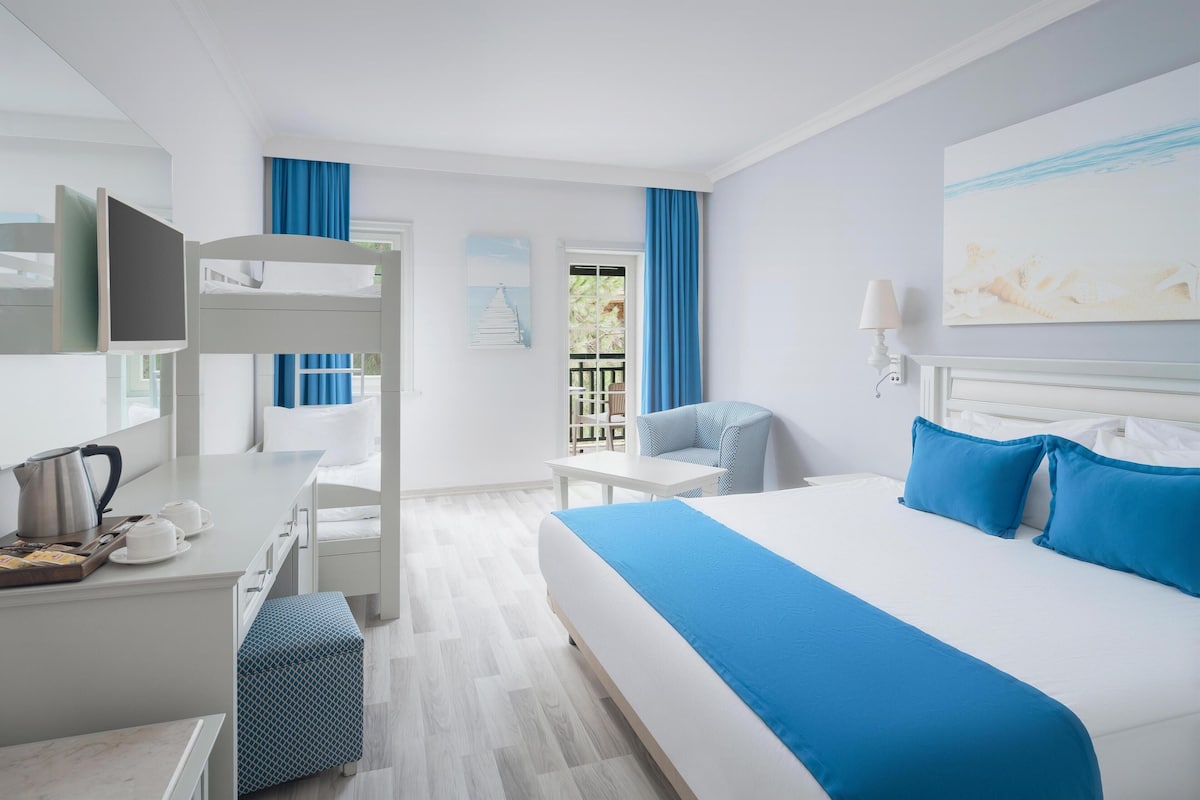 A hotel room with a bunk bed, a desk with a kettle, a chair, a bed with blue bedding, and a painting on the wall.