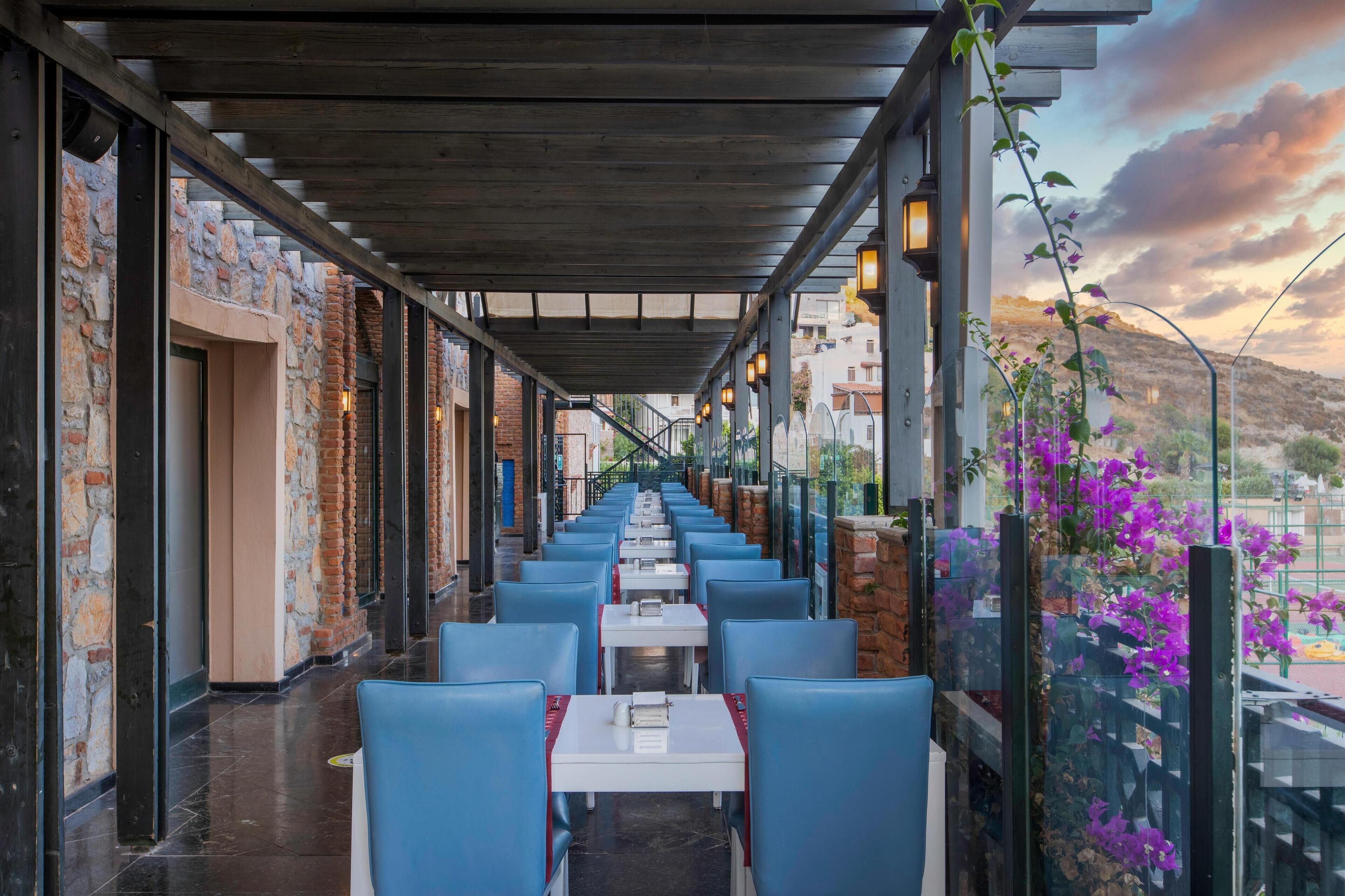 A covered outdoor seating area with blue chairs and tables, stone walls, and a view of the landscape.
