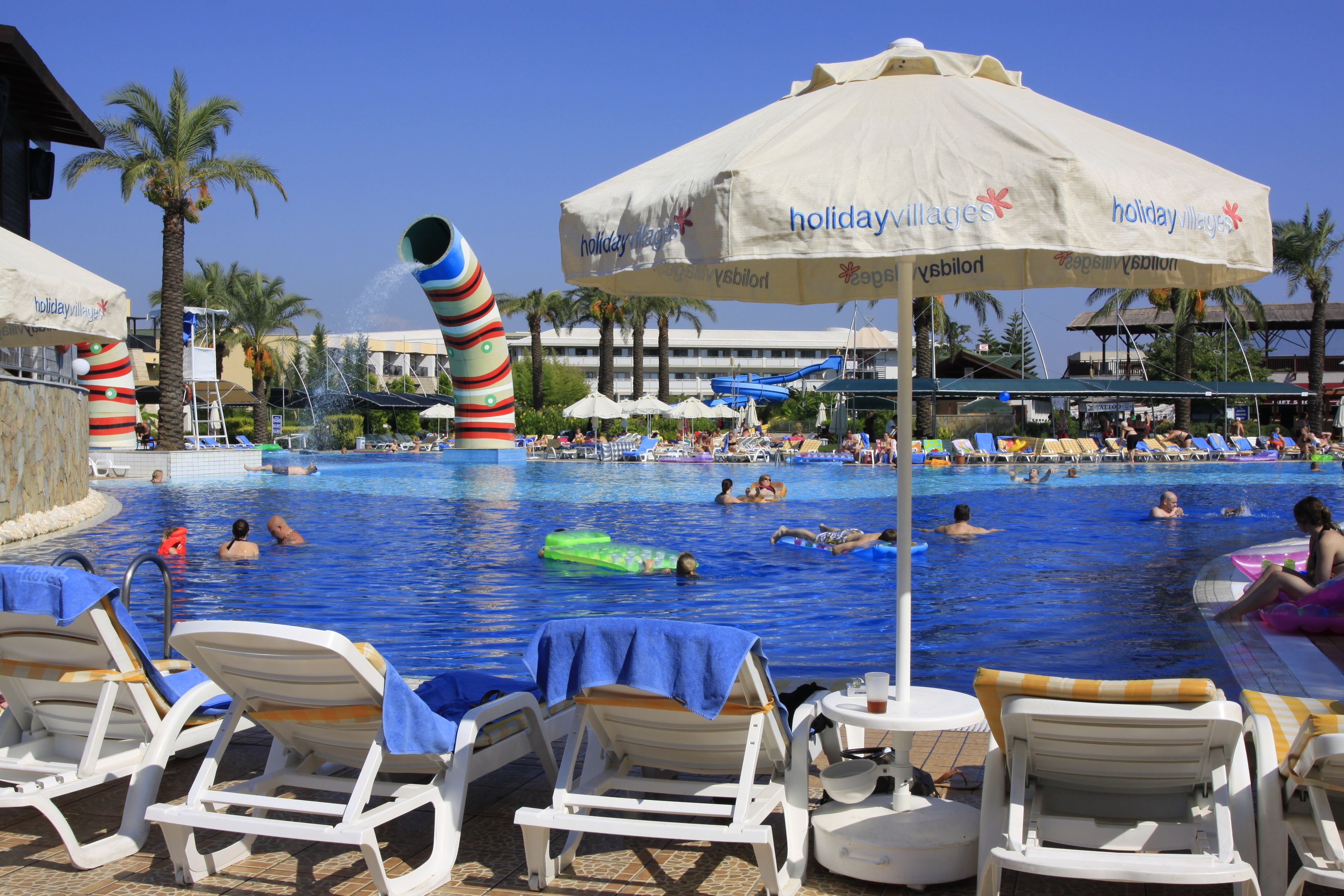 Un espace piscine agrémenté de chaises longues et de parasols, entouré de palmiers et surplombé d’un ciel d’un bleu limpide.