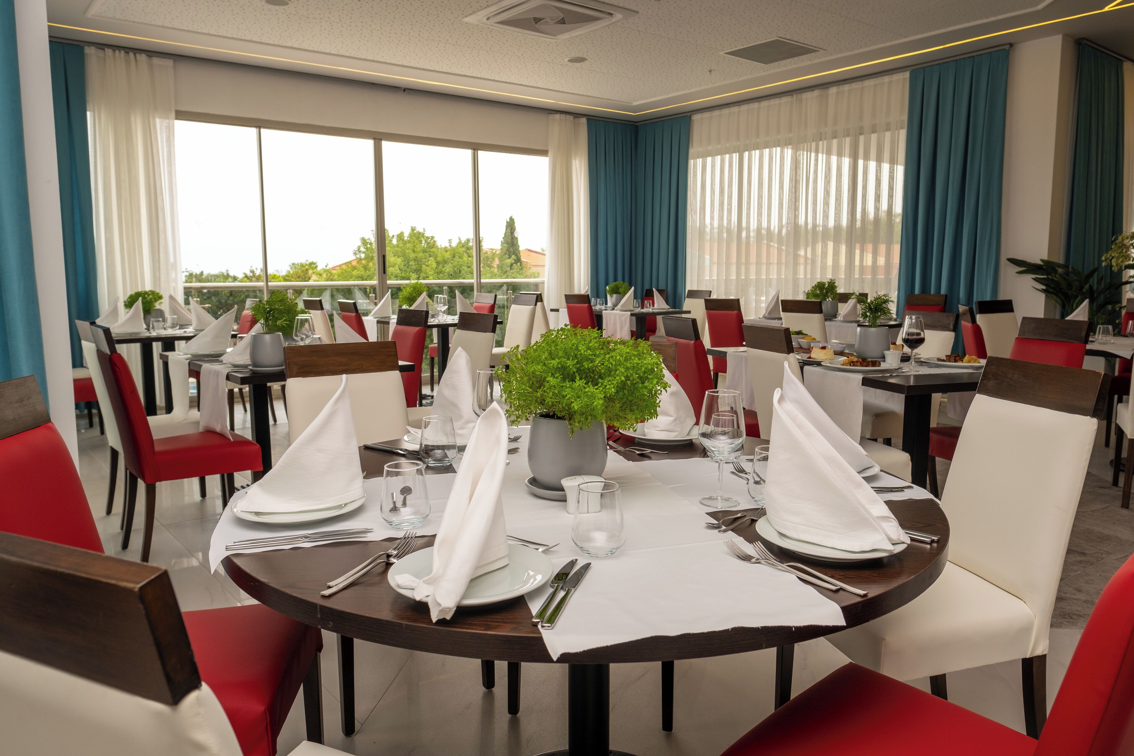 A dining area with round tables, red and white chairs, and potted plants.