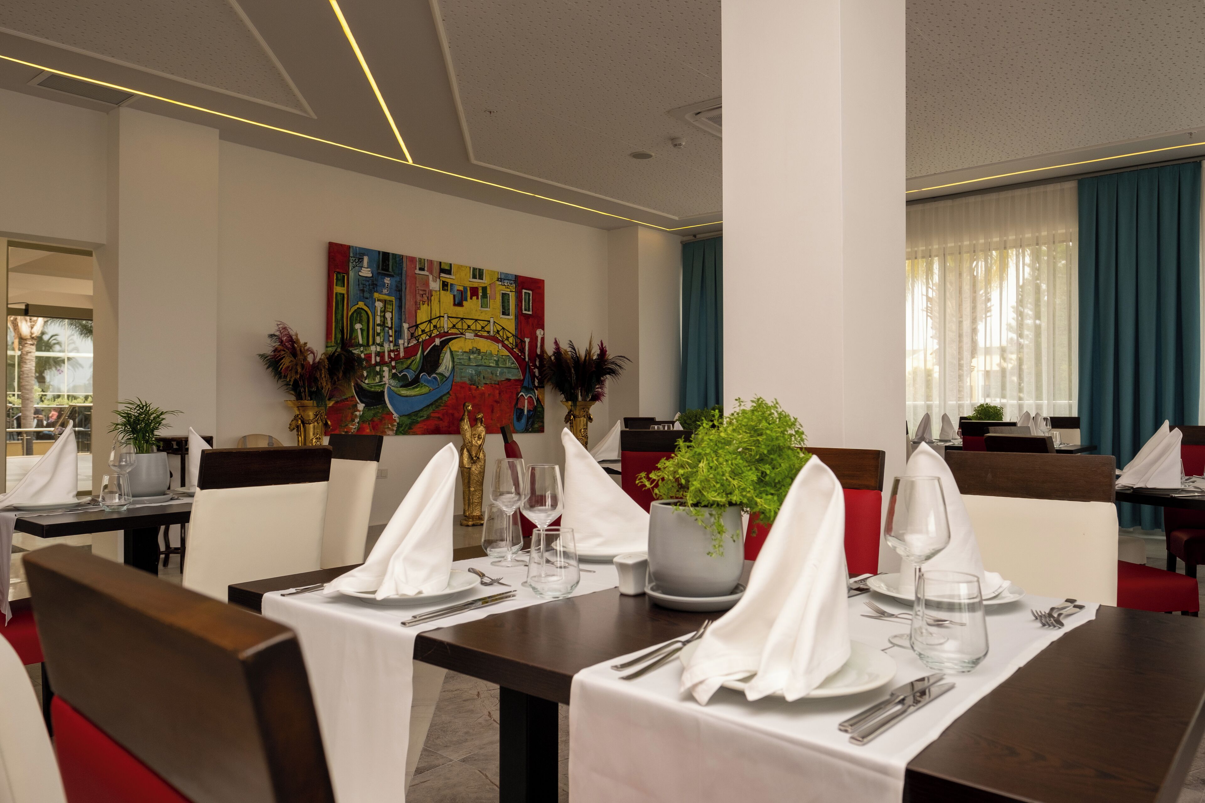 A dining area with tables set for two, featuring white tablecloths, silverware, and glassware. There is a colorful painting on the wall and a potted plant on the table.