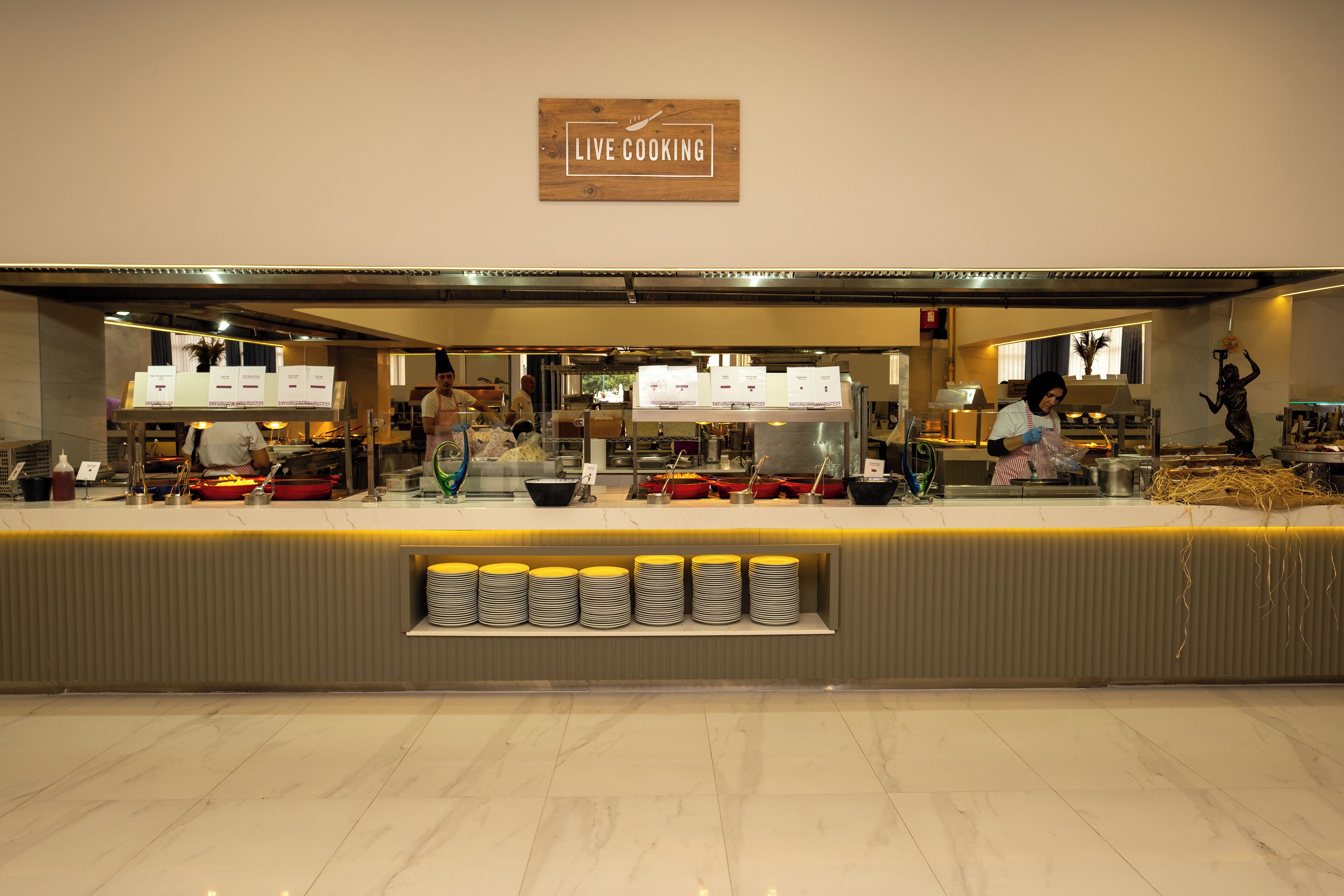 A restaurant with a 'Live Cooking' sign, a counter with stacked plates, and kitchen staff preparing food.
