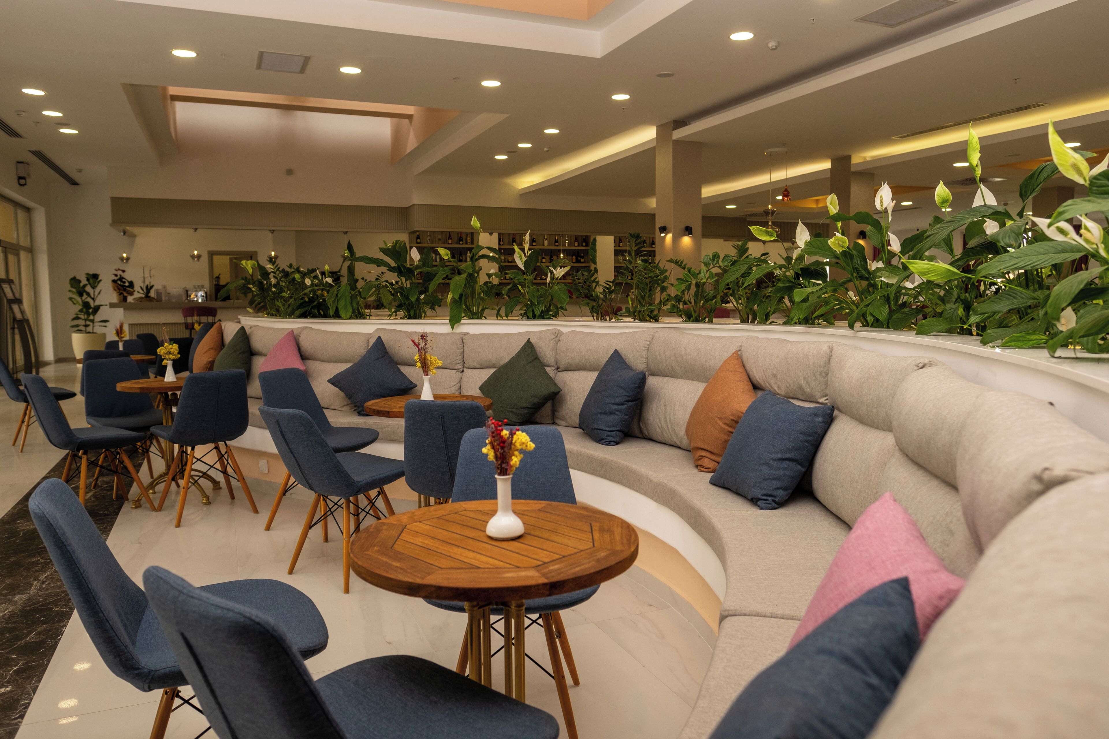 Een moderne zithoek met een gebogen bank, blauwe stoelen, een ronde houten tafel en diverse planten.