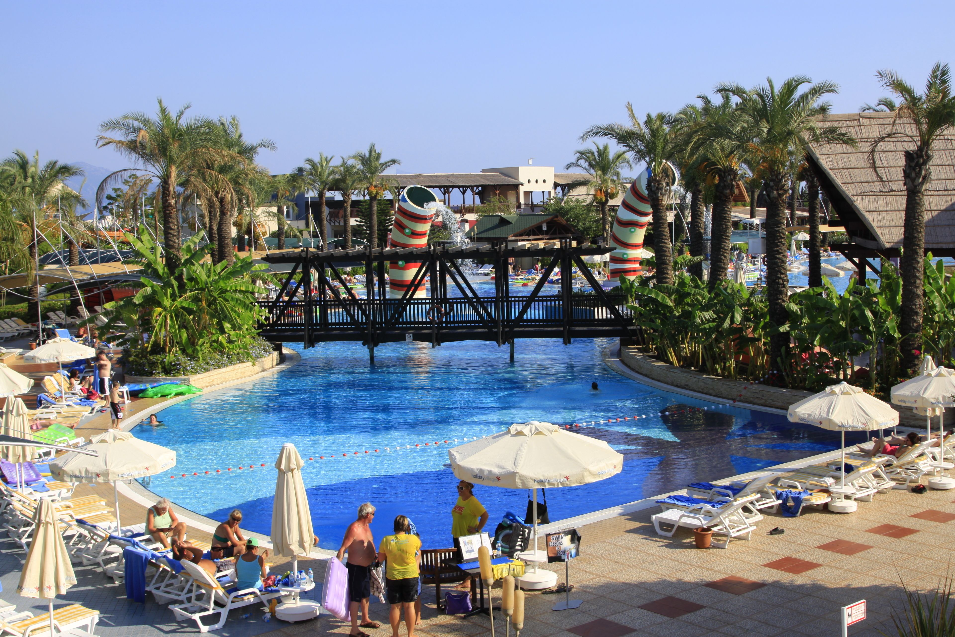 A swimming pool area with lounge chairs, umbrellas, and palm trees.
