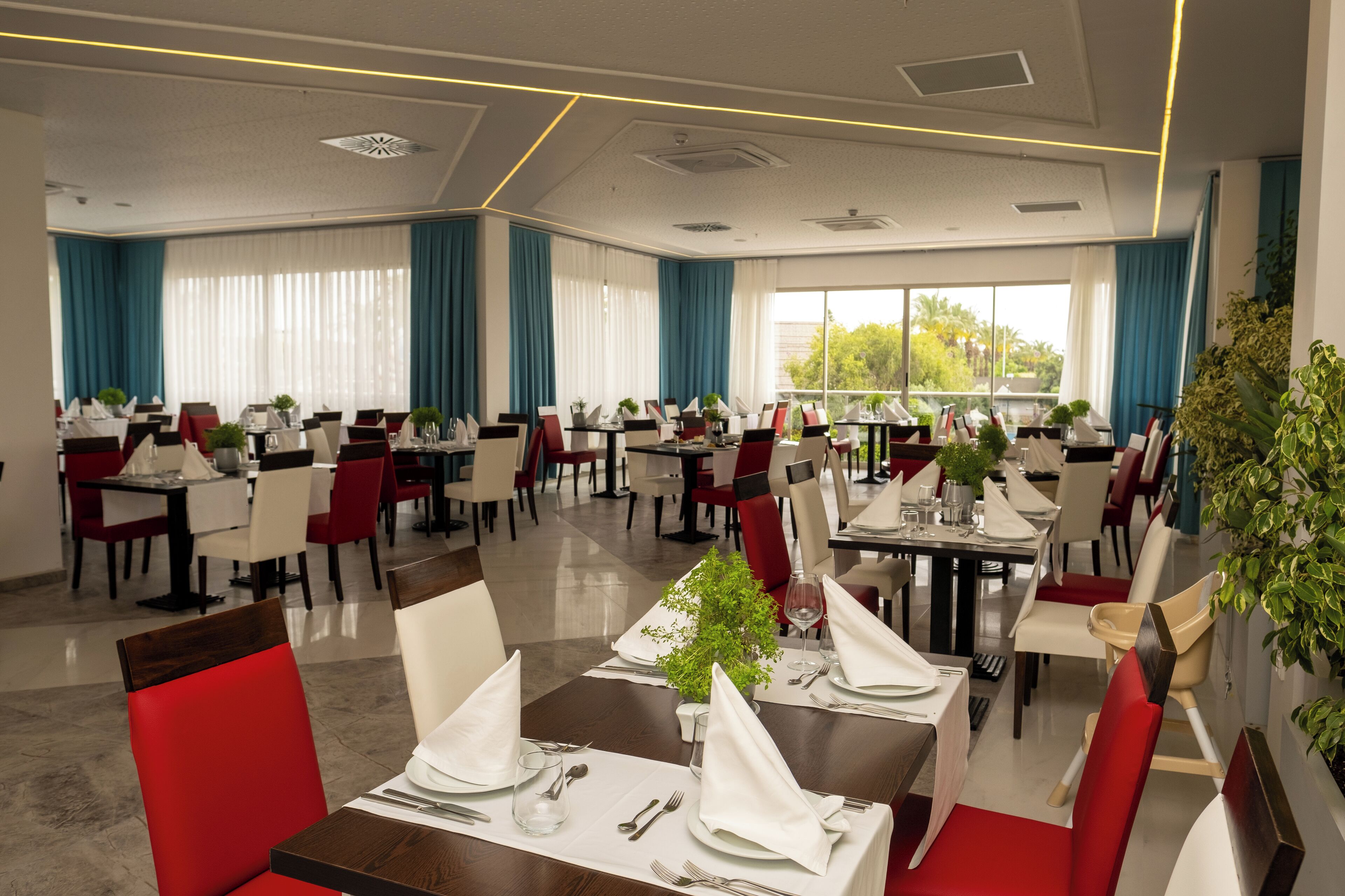Un comedor amplio con varias mesas y sillas, ventanas grandes con cortinas y vistas a zonas verdes.
