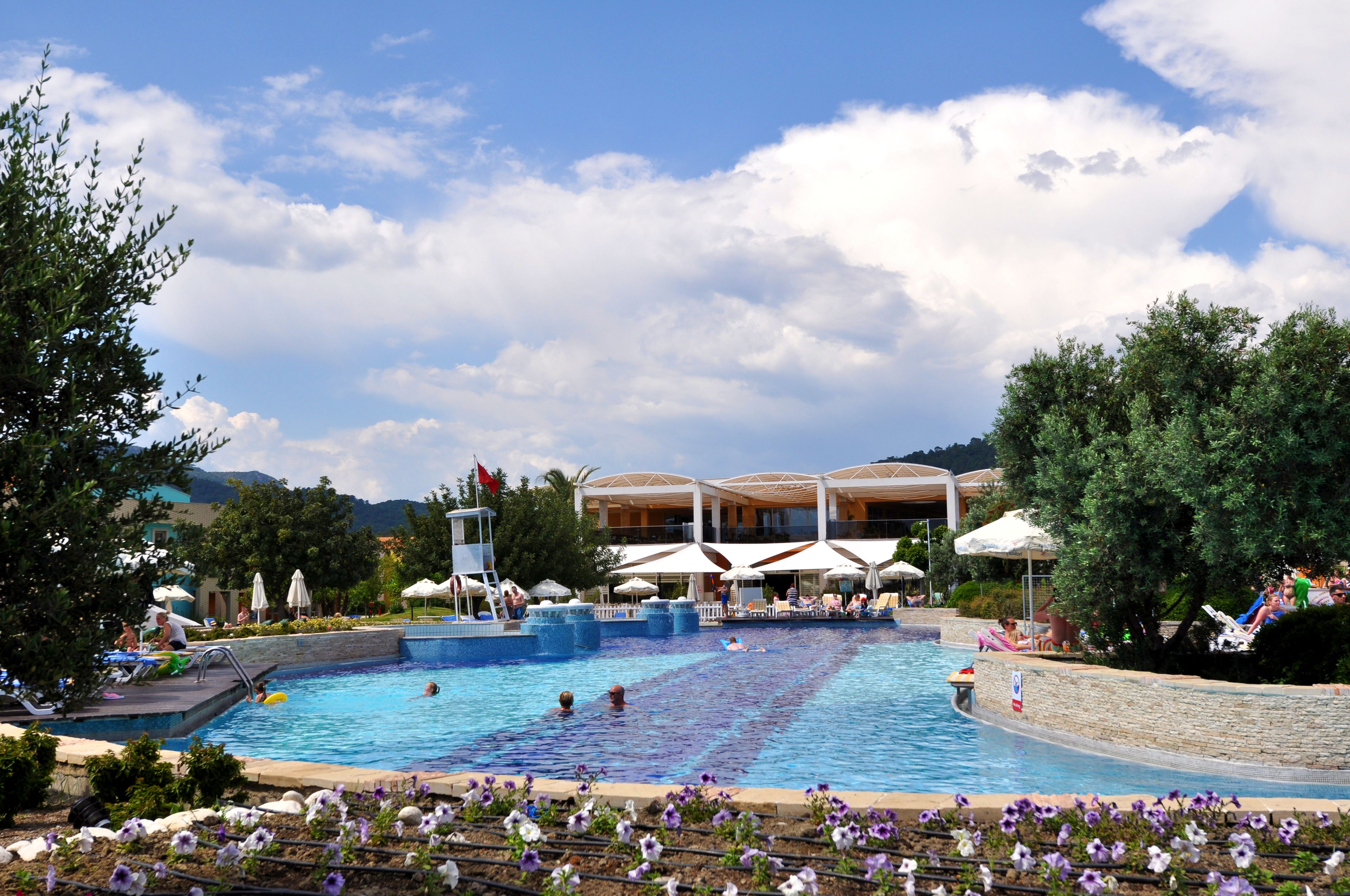 A large outdoor swimming pool with lounge chairs and umbrellas, surrounded by flowers and trees.
