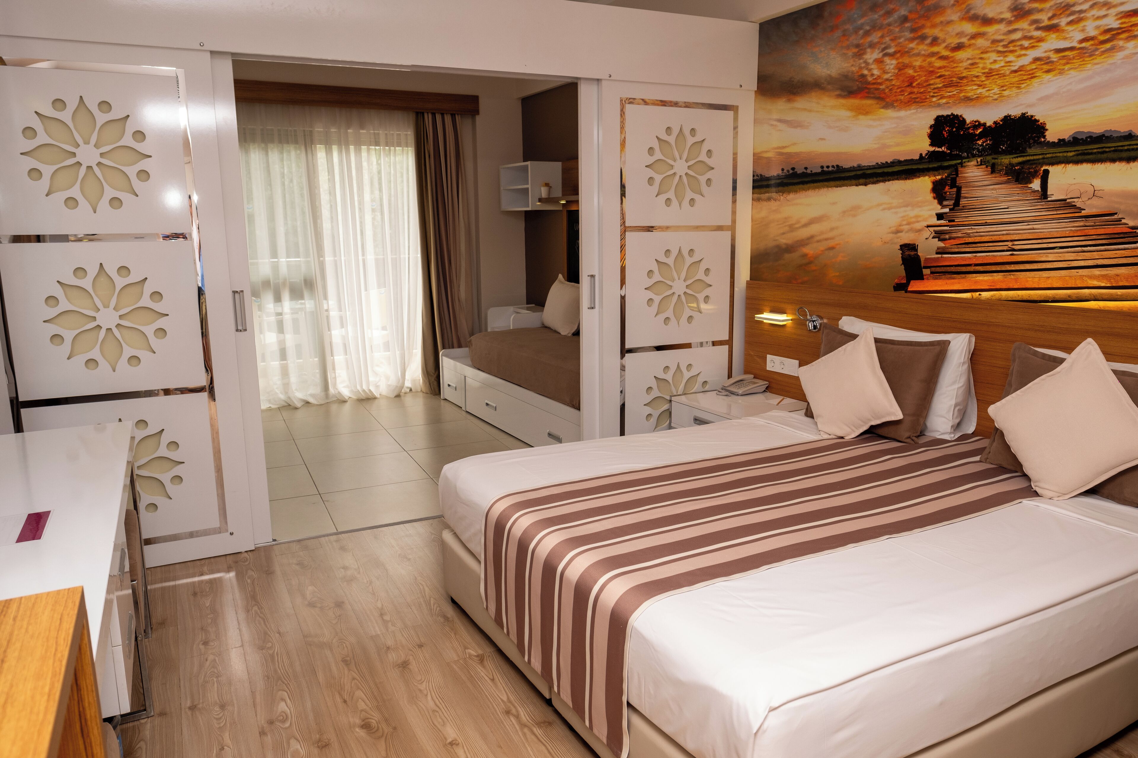 A hotel room with a bed, a desk, and a painting of a sunset over a pier.