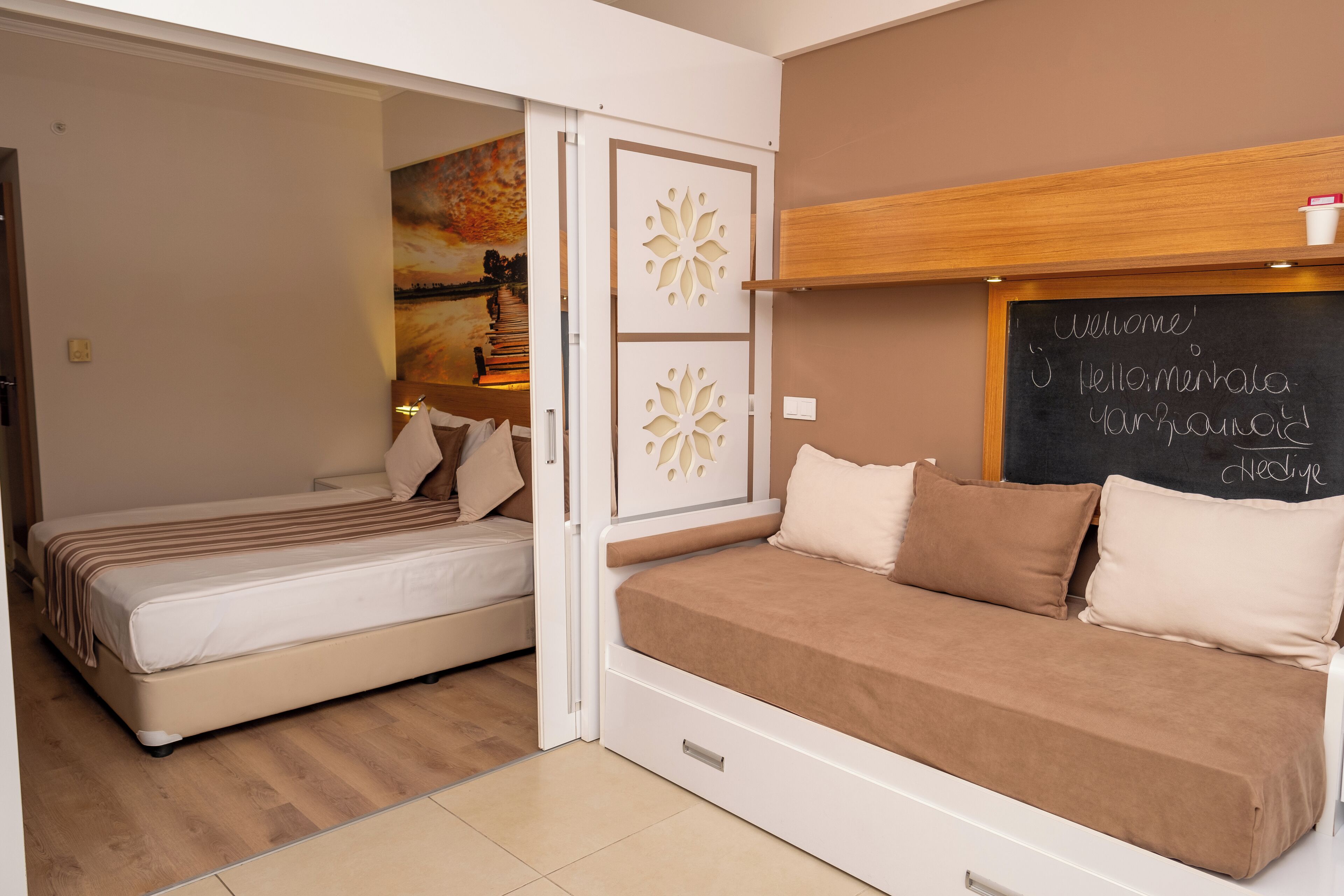 A bedroom with two beds, a wooden headboard, a chalkboard with 'Welcome' written on it, and a painting on the wall.