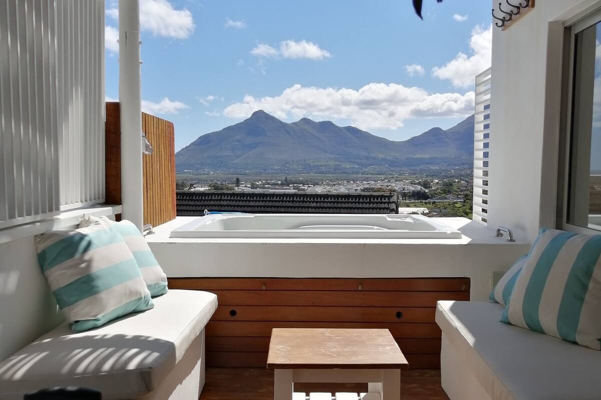 A balcony with a wooden table, white cushioned seating, and a view of a mountain.