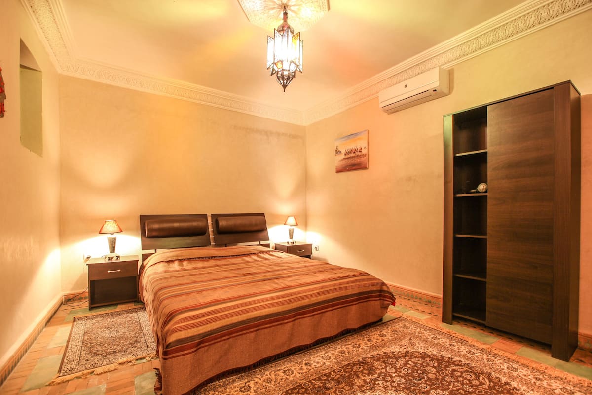 A bedroom with a large bed, two bedside tables with lamps, a wardrobe, and a chandelier.