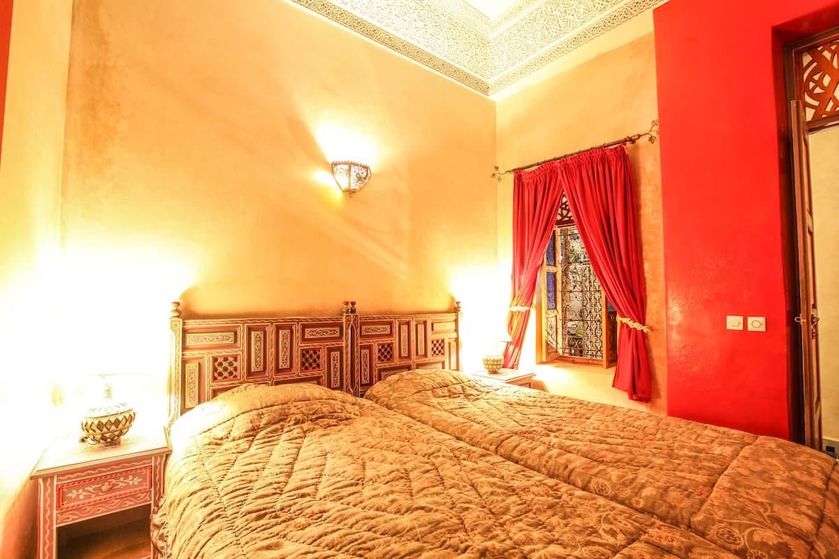 A room with a large bed, a nightstand with a lamp, and a window with red curtains.