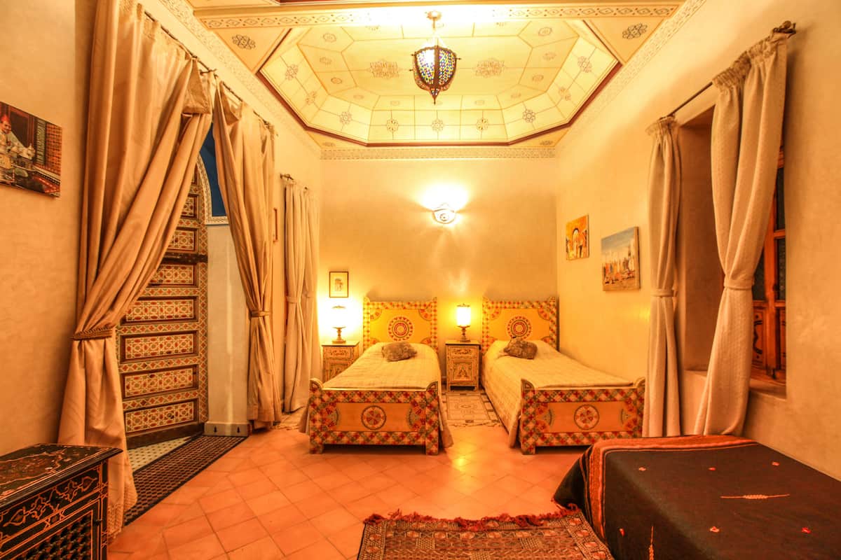 A room with two beds, a chandelier, patterned walls, and a staircase.