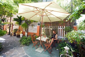 Un patio con una mesa y sillas bajo un paraguas grande, rodeado de plantas en macetas y una cerca blanca.