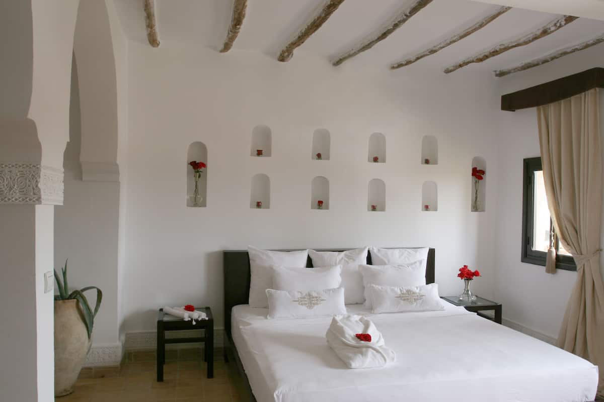 A neatly arranged bedroom with a large bed, multiple pillows, a bedside table with a vase of flowers, and a potted plant.