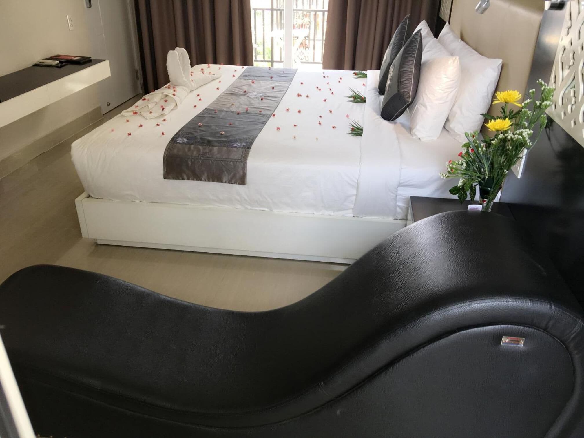 Habitación de hotel moderna con una cama grande, una silla de masaje negra y una pequeña mesa con un jarrón de flores.