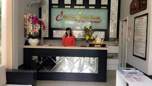 A hotel reception desk with a large clock displaying times for various cities, a receptionist, and a decorative wall with flowers and a slogan. - Mui Ne Sports Hotel (Mui Ne)