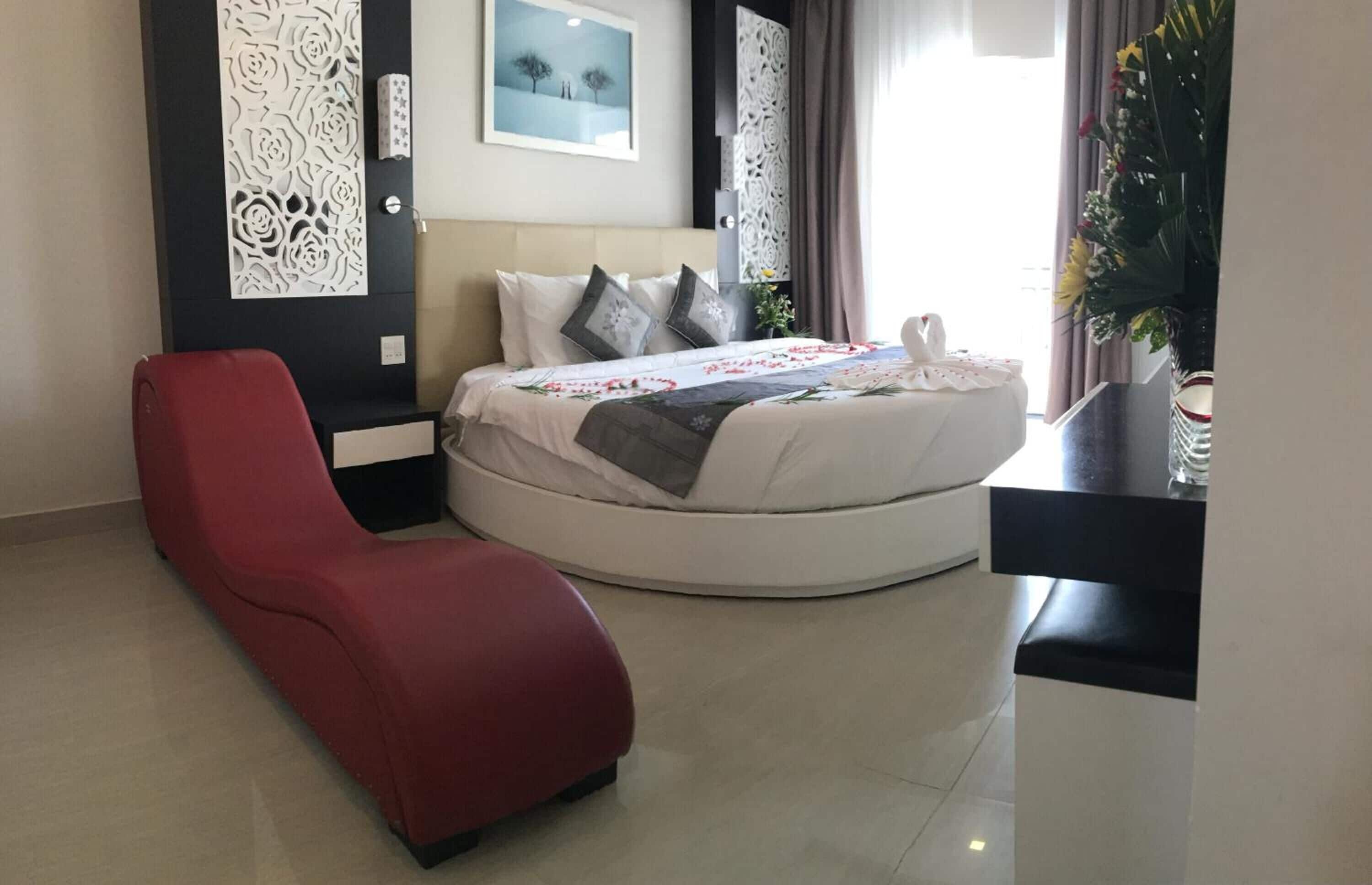 A modern hotel room with a round bed, a red lounge chair, decorative wall panels, and a large window with curtains.