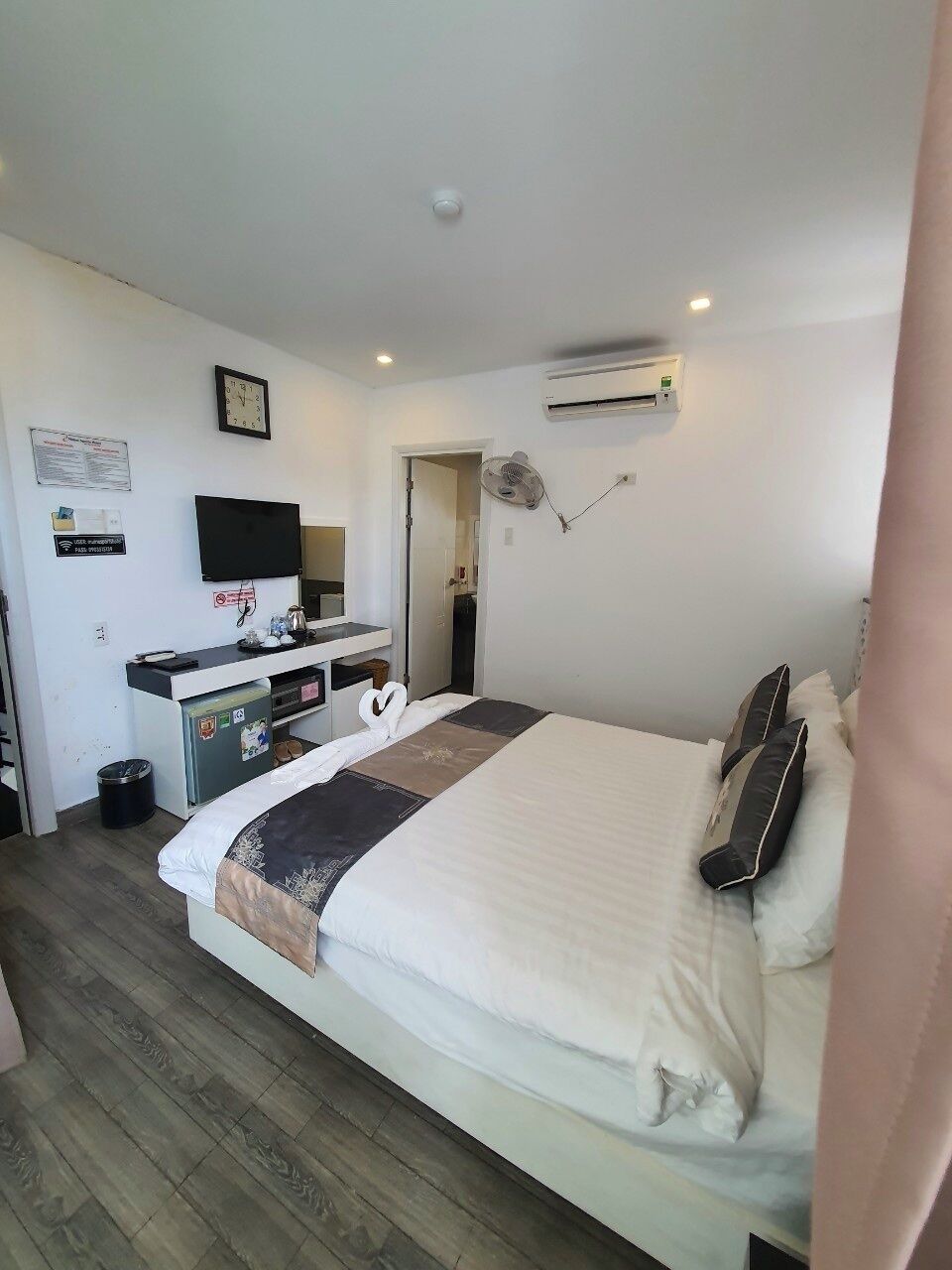 A hotel room with a bed, a desk with a flat-screen TV, an air conditioning unit, and a wall-mounted fan.