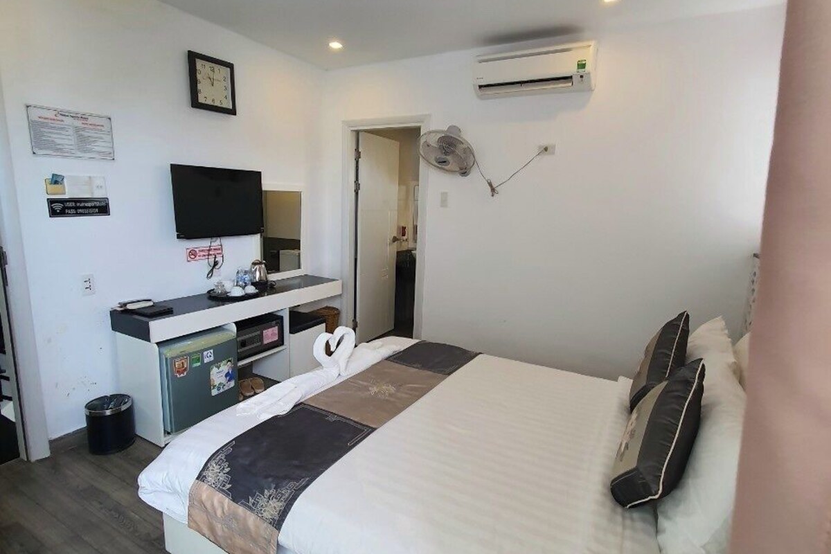 Una habitación de hotel con cama, escritorio con televisor de pantalla plana, aire acondicionado y ventilador de pared.