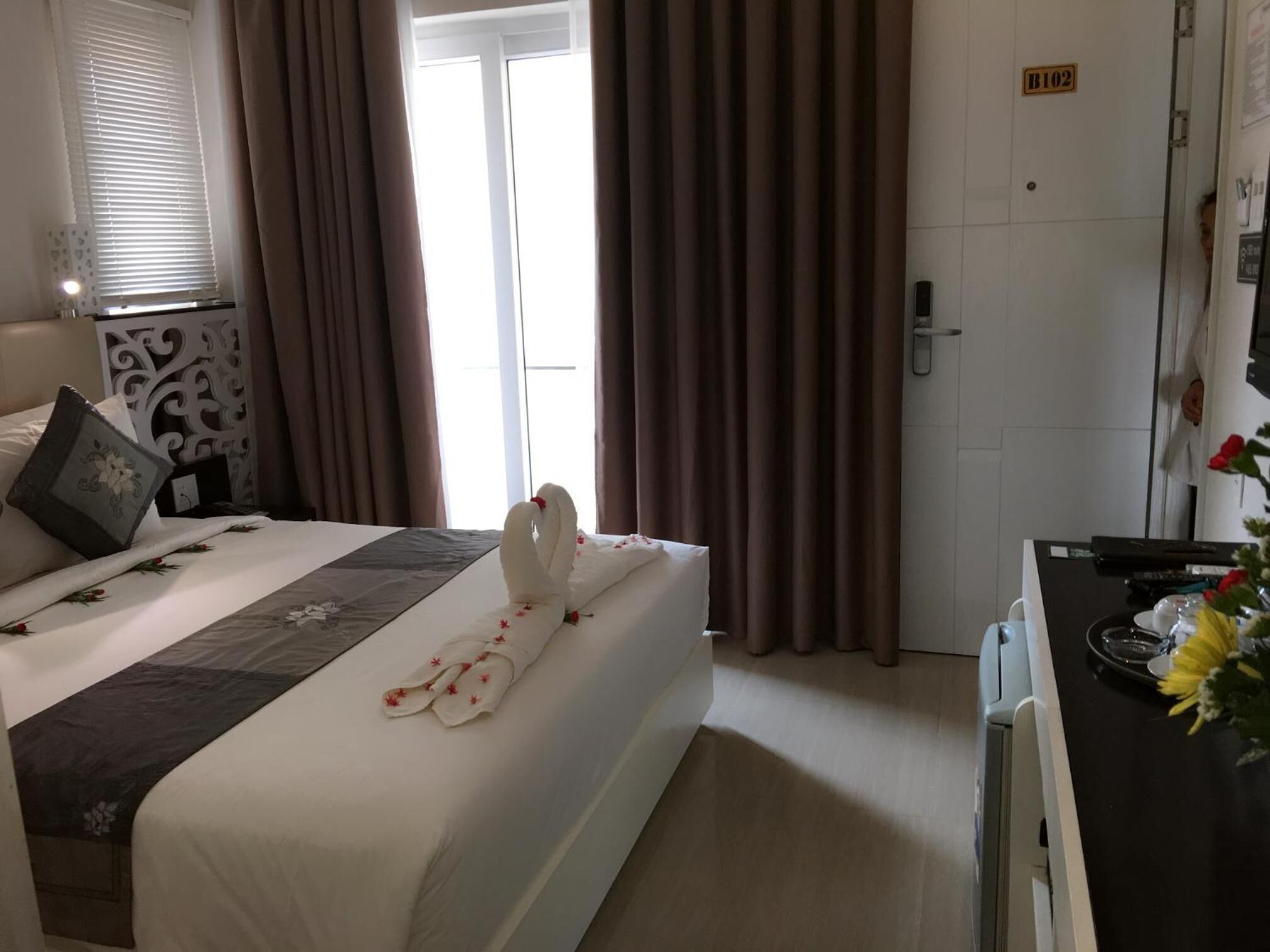A hotel room with a neatly made bed, a desk with a vase of flowers, and a door with a sign.