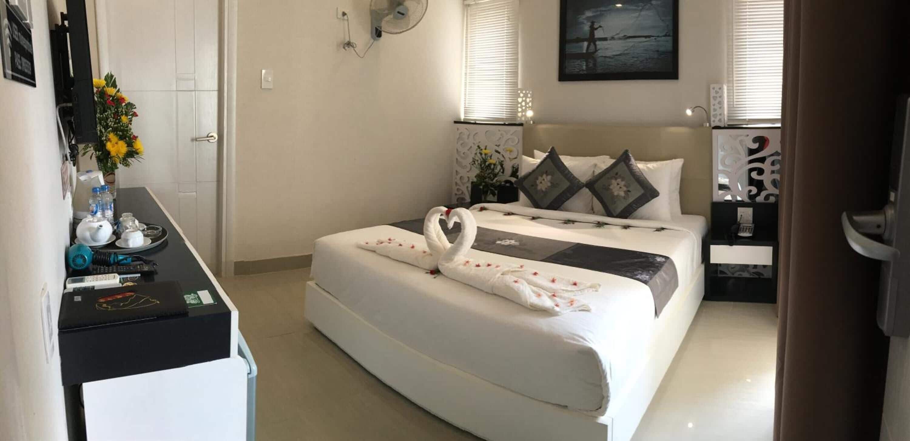 Une chambre d’hôtel avec un lit, un bureau avec des articles de toilette, un ventilateur fixé au mur et un tableau.