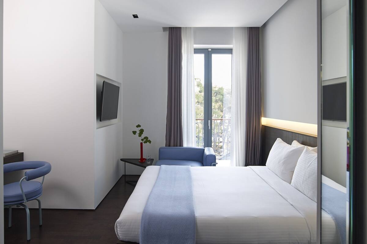 A modern hotel room with a large bed, a seating area with a chair, a small table with a plant, and a balcony view.
